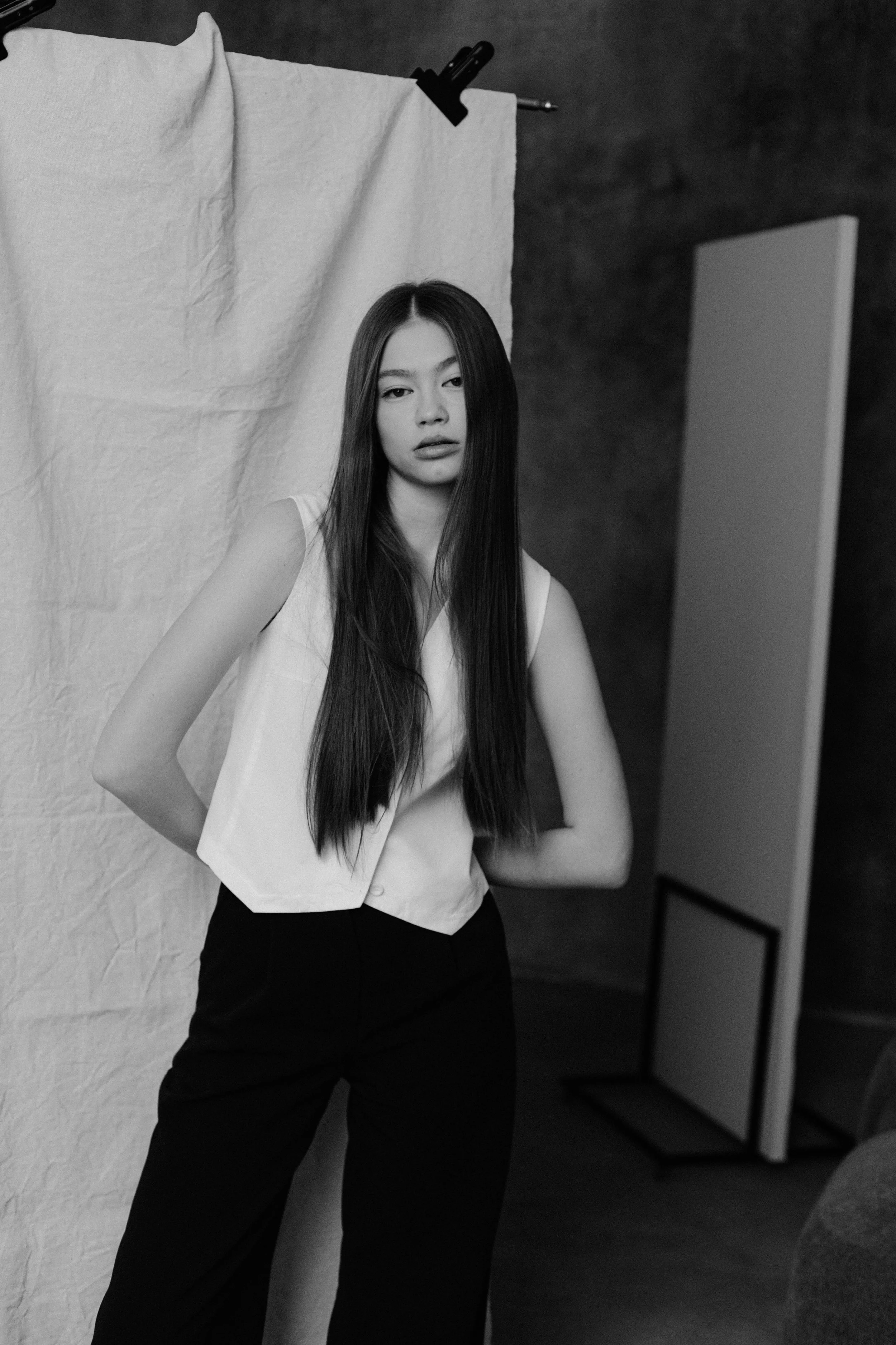 Black and white photo of a woman with long dark hair standing in front of a backdrop, wearing a sleeveless top and black pants, with a neutral expression.