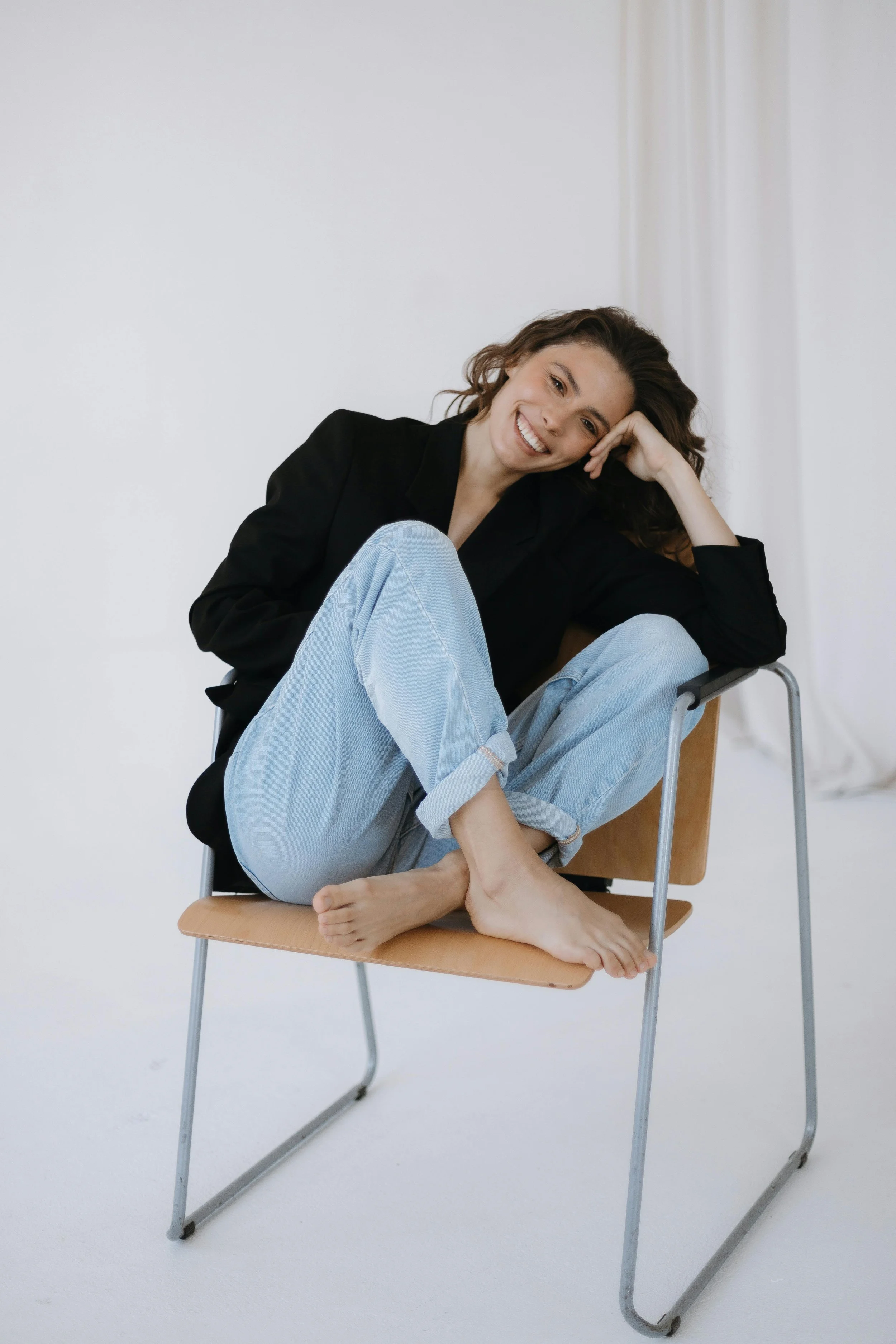 A woman sitting on a wooden chair with metal legs, smiling and relaxing with her legs curled up, wearing a black blazer and light blue jeans, in a bright studio setting.