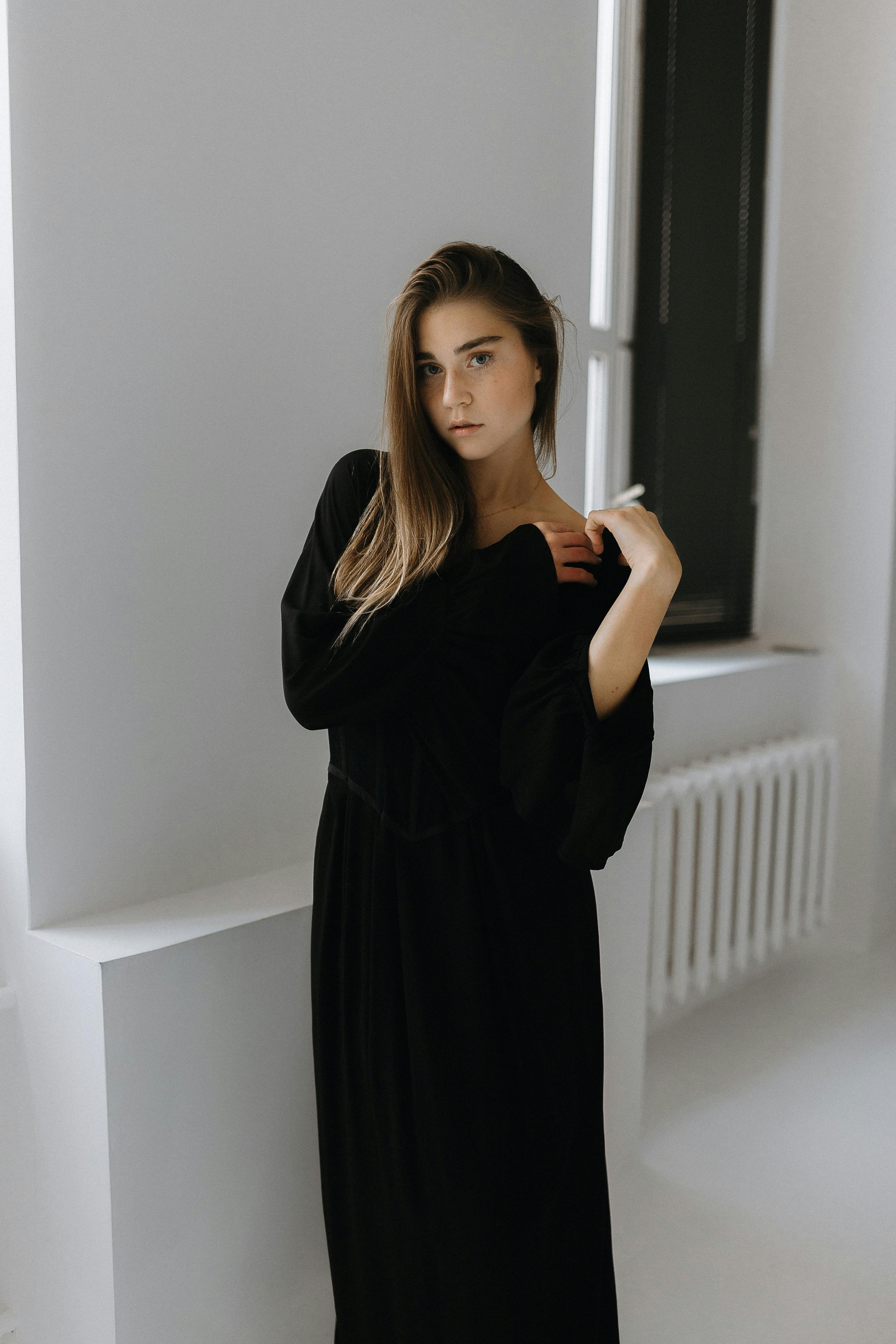 A young woman with long, brown hair and blue eyes, wearing a black dress, standing indoors near a window with black shutters, in front of a white wall and radiator.