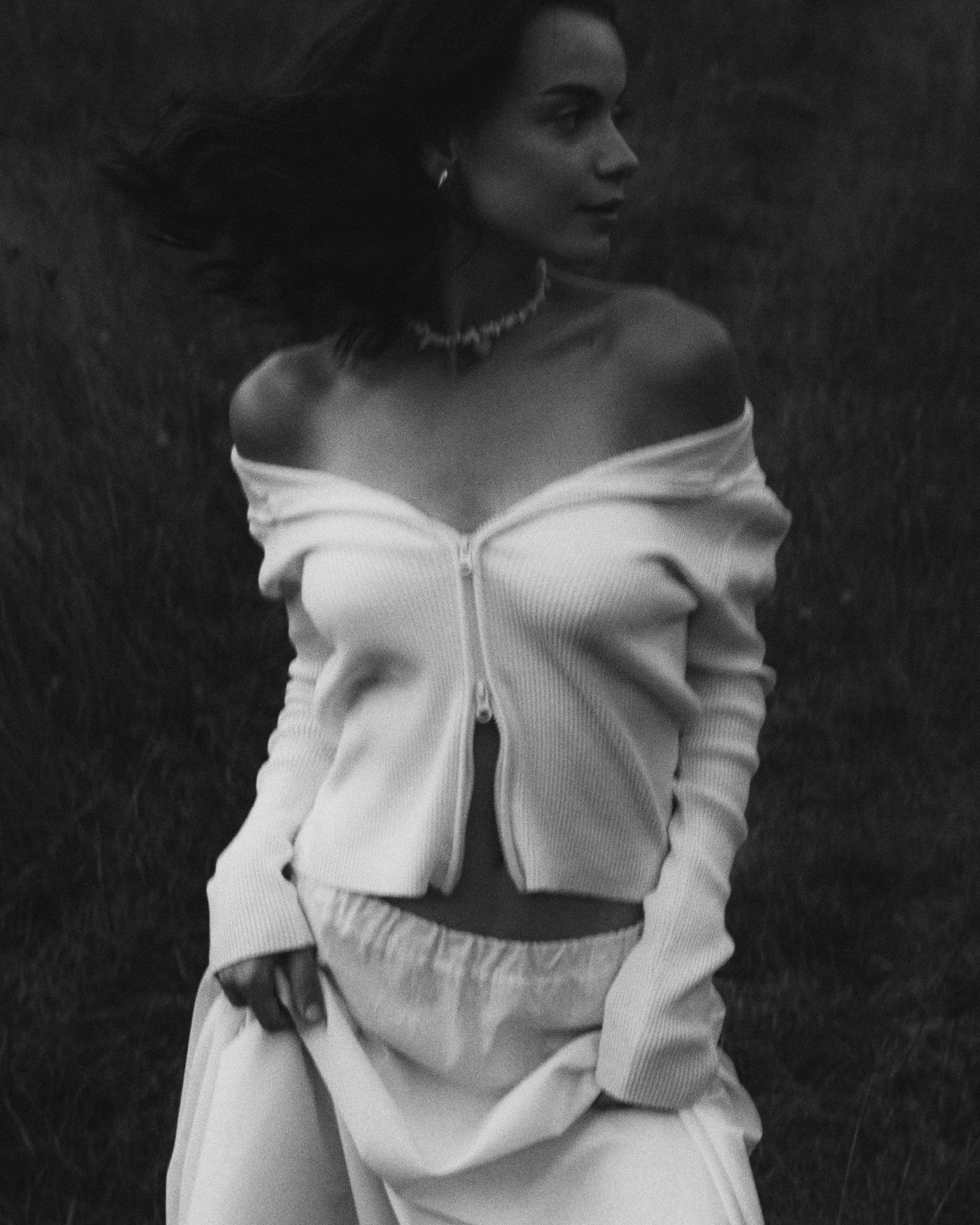 Black and white photo of a woman with long hair, wearing an off-shoulder zip-up top and pants, standing outdoors on a grassy field.