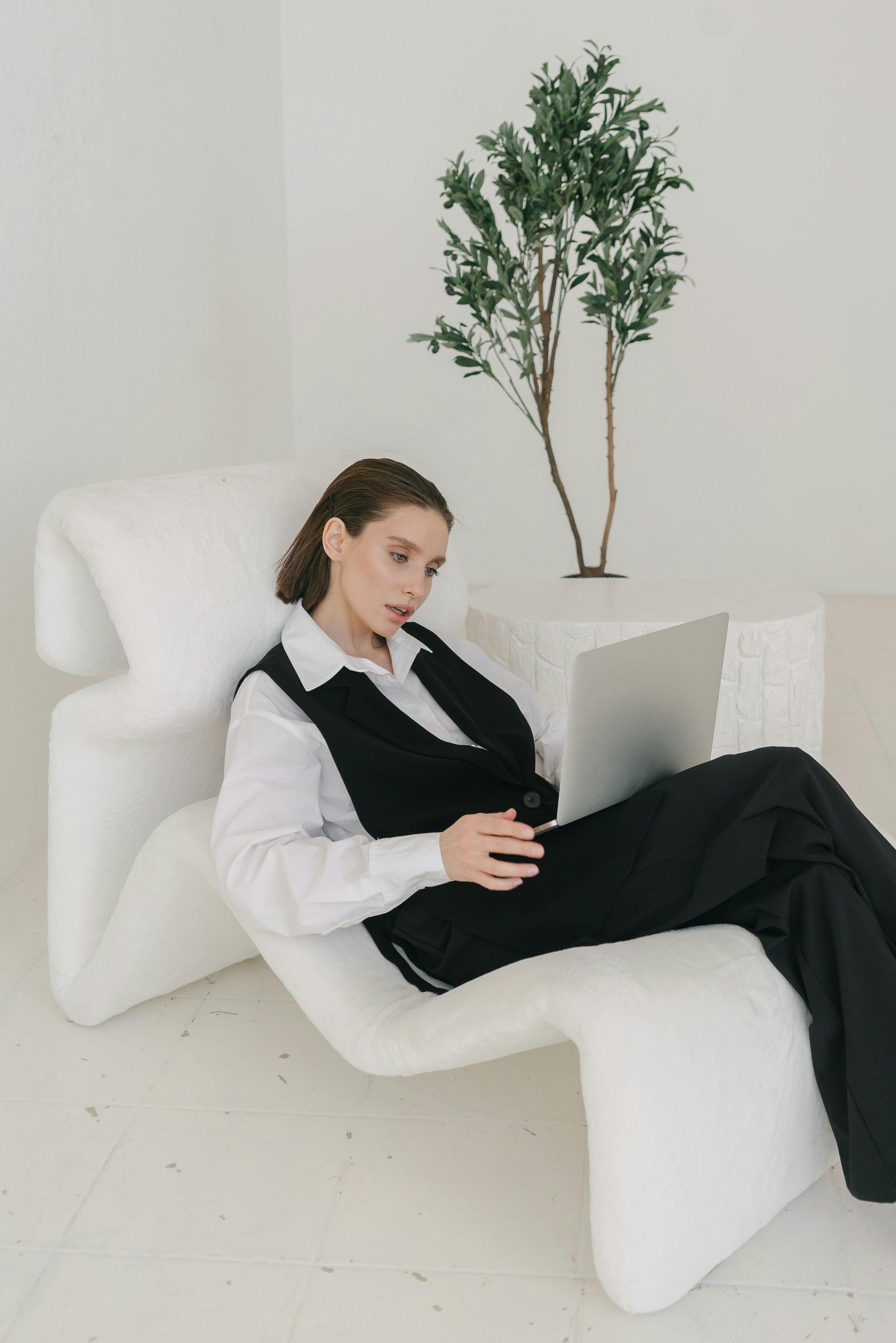 A woman sitting on a white modern lounge chair, working on a laptop, in a minimalist room with a large potted plant in the background.