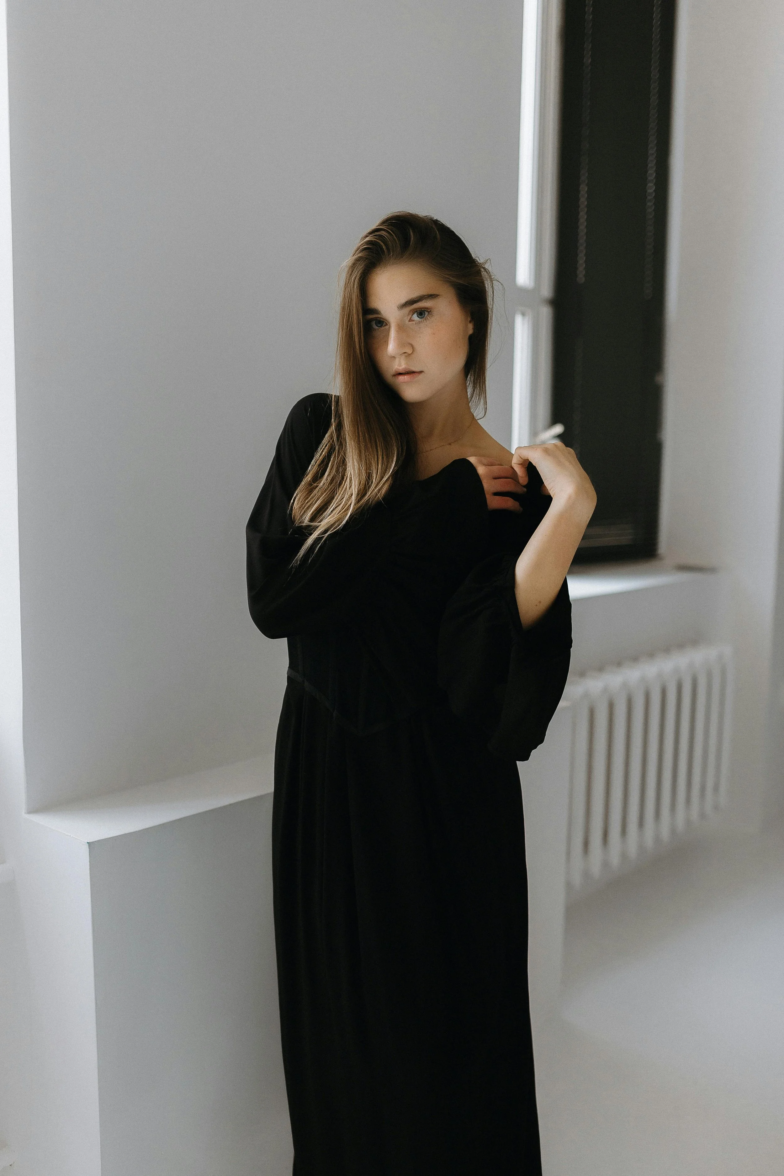 A young woman with long light brown hair, wearing a black dress, standing near a window in a minimalist room with white walls and a radiator.