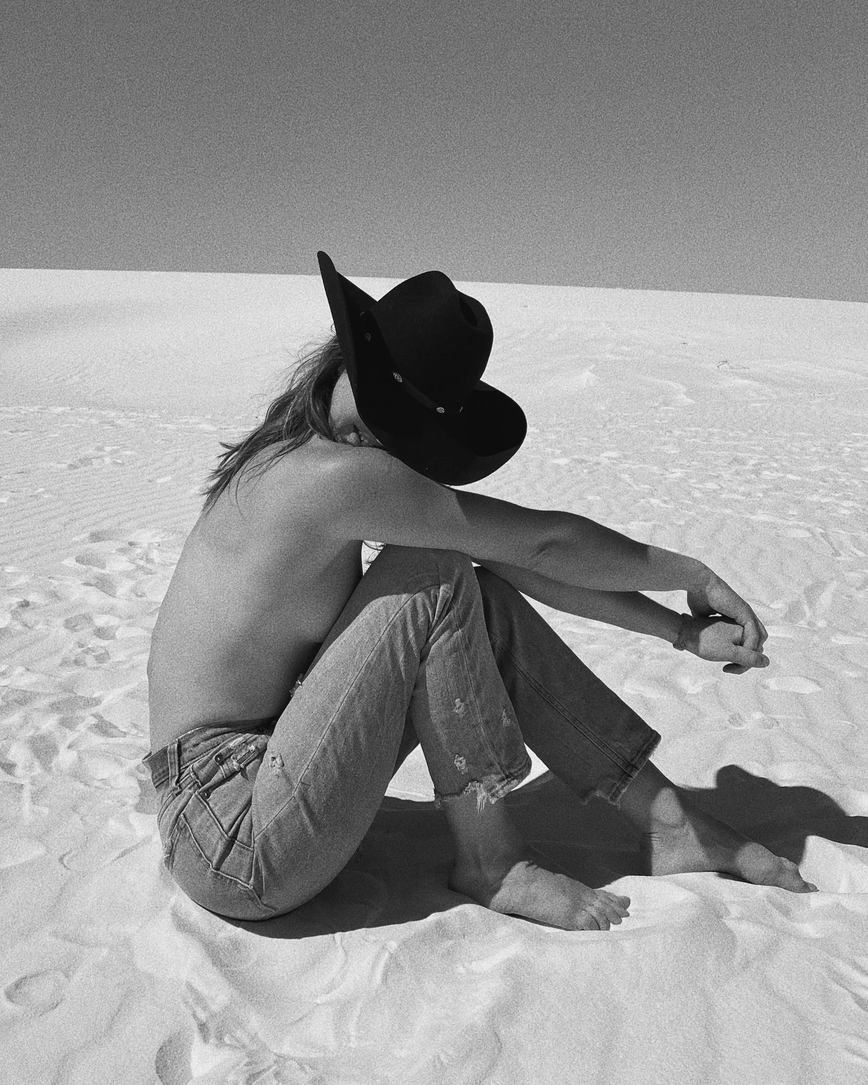 A person sitting on the sandy ground of a desert or beach, wearing a large black hat, with their back to the camera, shirtless, with torn jeans, and barefoot.