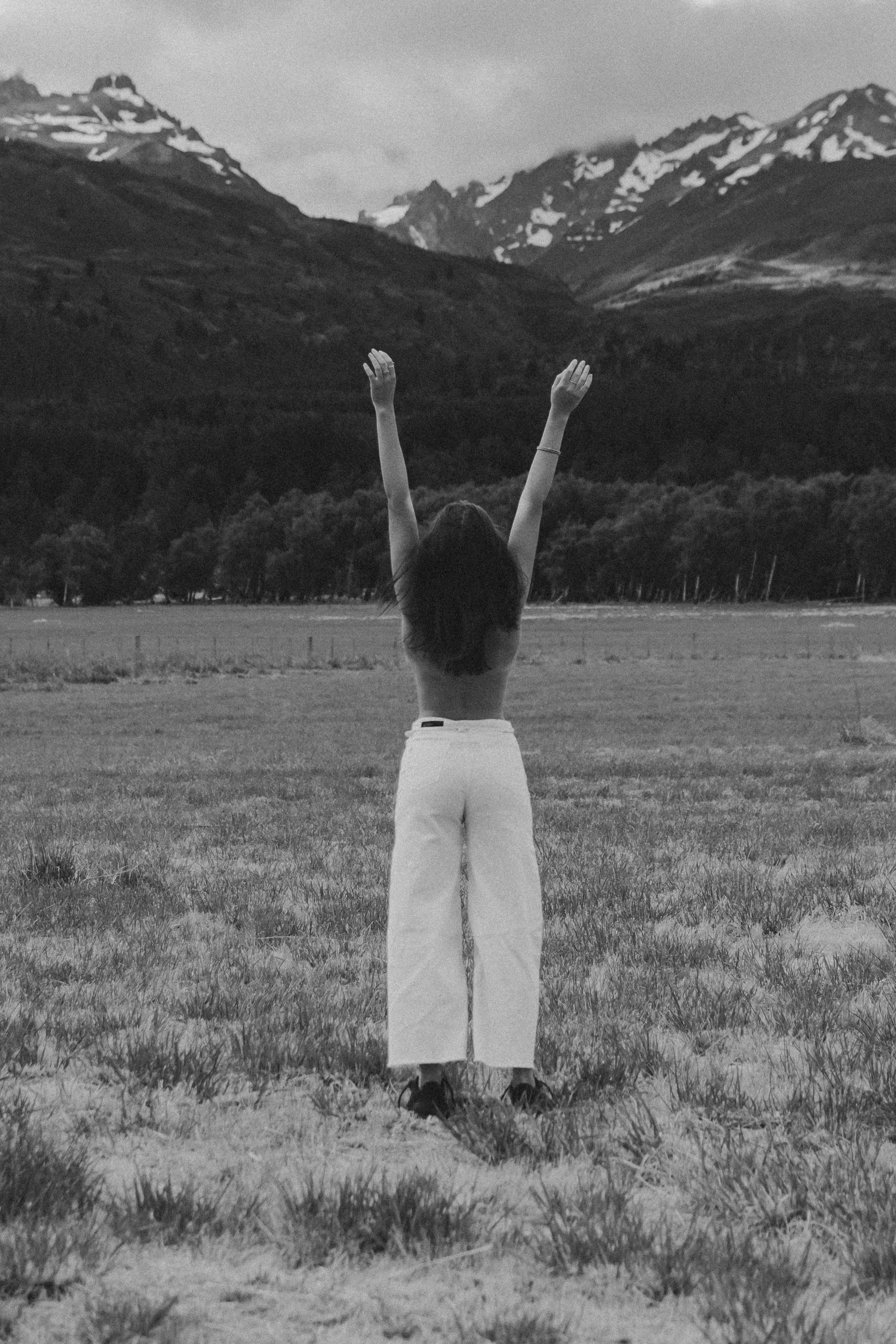 A woman standing in a grassy field with her arms raised, facing mountains with snow patches in the background, in black and white.