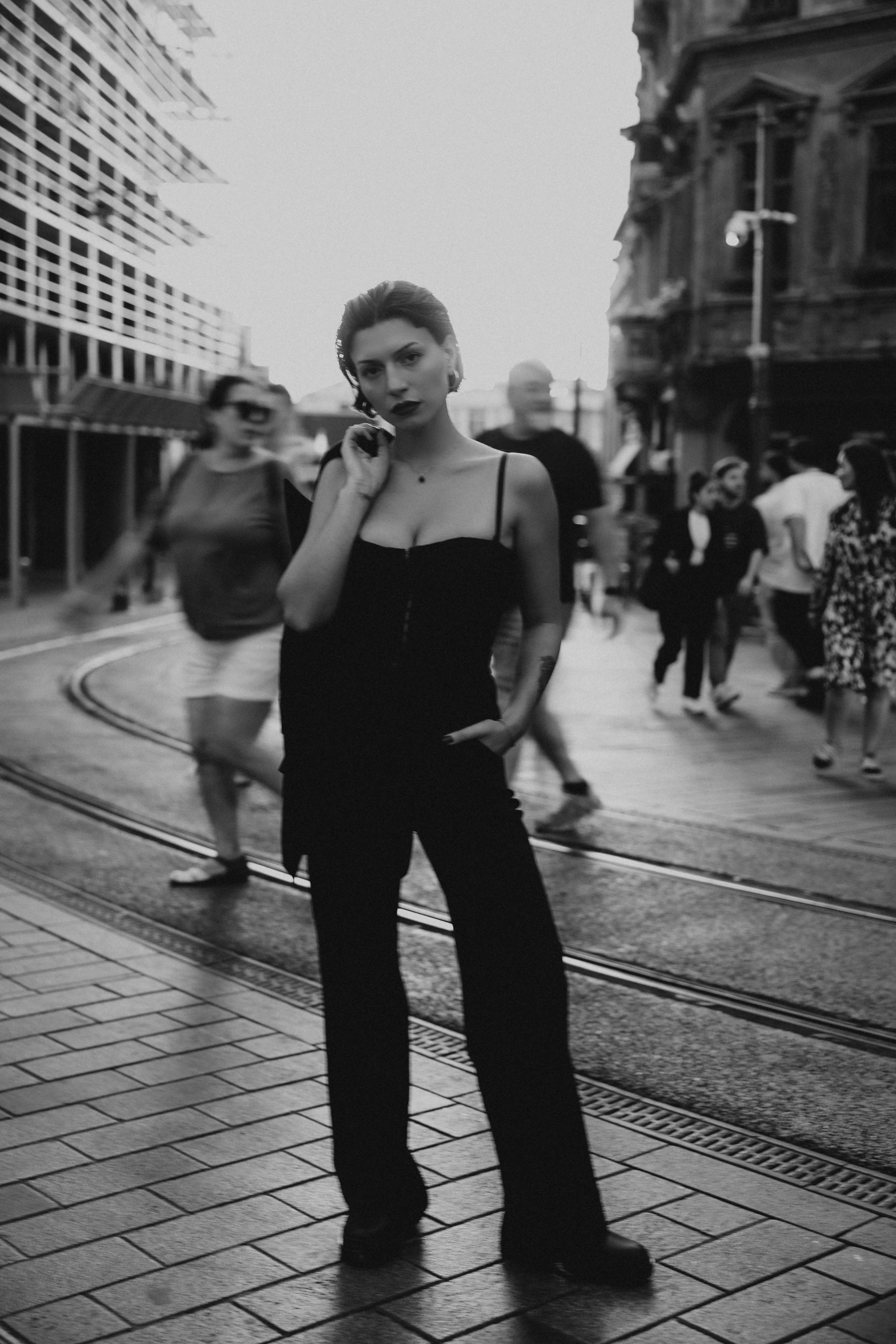 Black and white photo of a woman in a spaghetti strap top and wide-leg pants standing on a city street with tram tracks, with blurred pedestrians in the background.