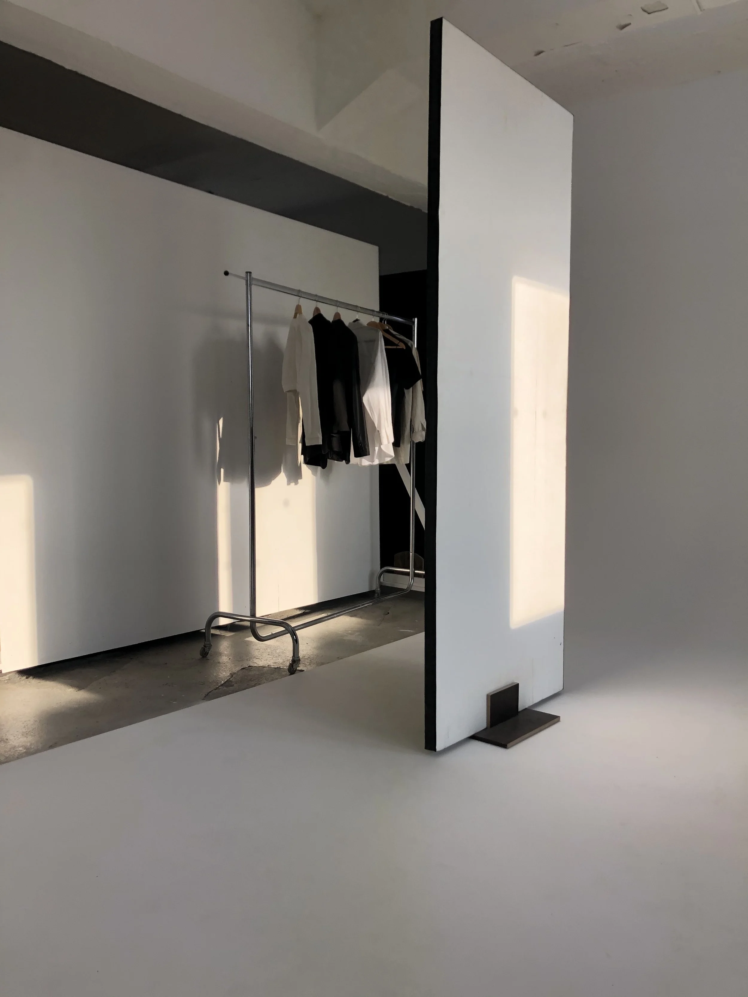 Clothing rack with white, black, and beige shirts hanging inside a minimalistic studio with white walls and a shadow of sunlight