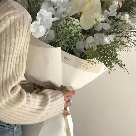 Person in a beige sweater holding a bouquet of white and green flowers wrapped in white paper.