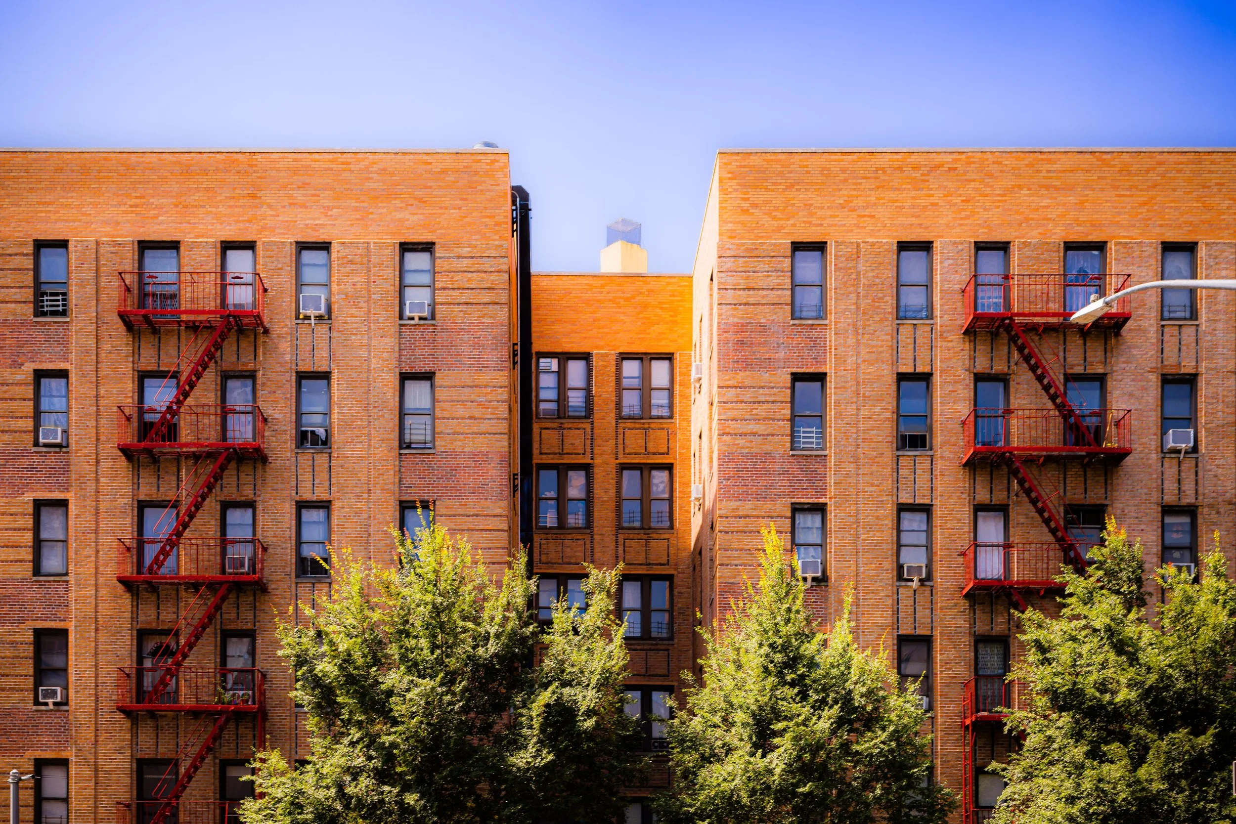 Two brick apartment buildings with fire escape staircases on the left and right sides, trees in the foreground, and a clear blue sky above.