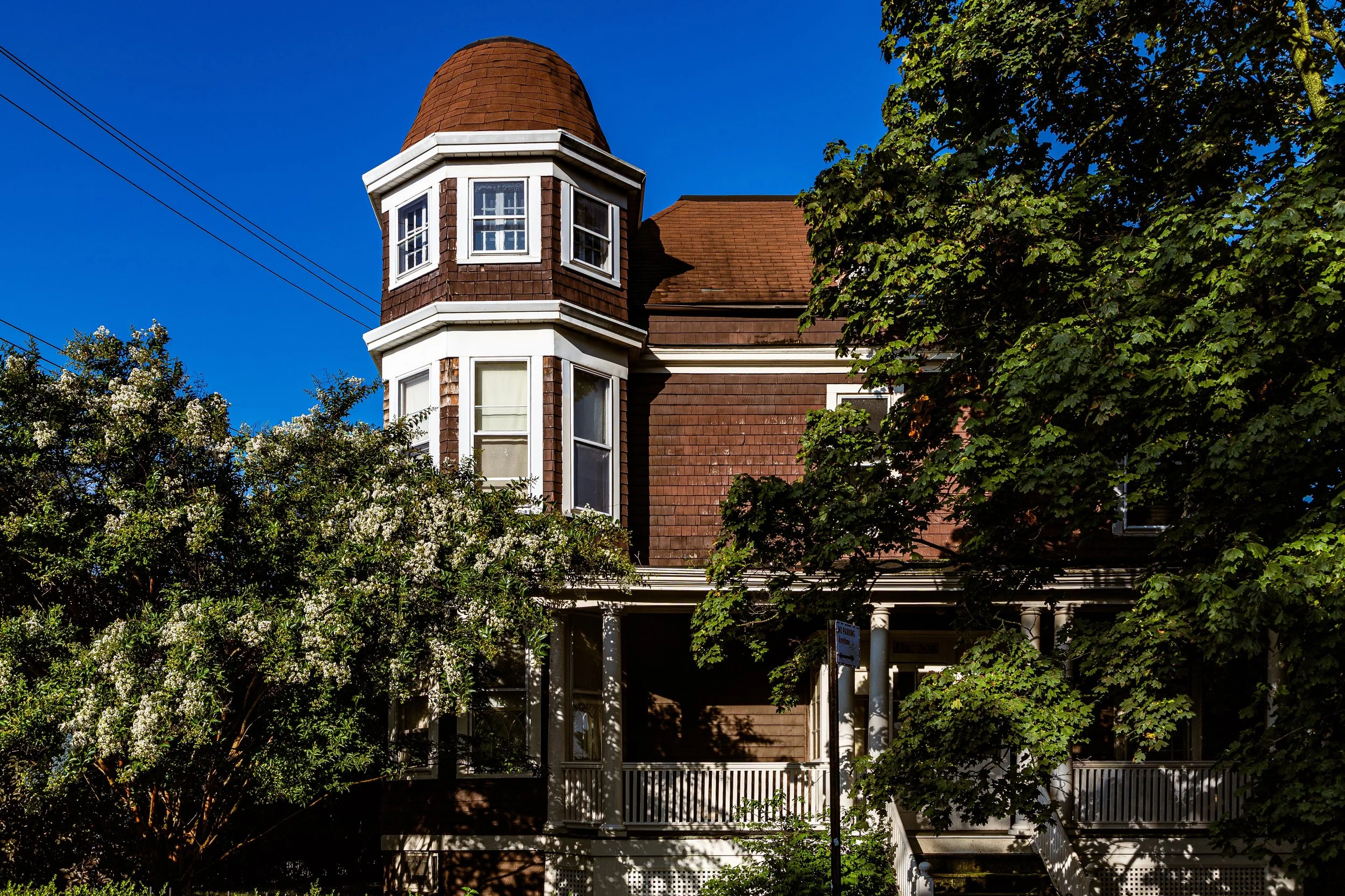 Old Victorian-style house with a rounded tower, brown shingles, white trim, and surrounded by trees under a clear blue sky.