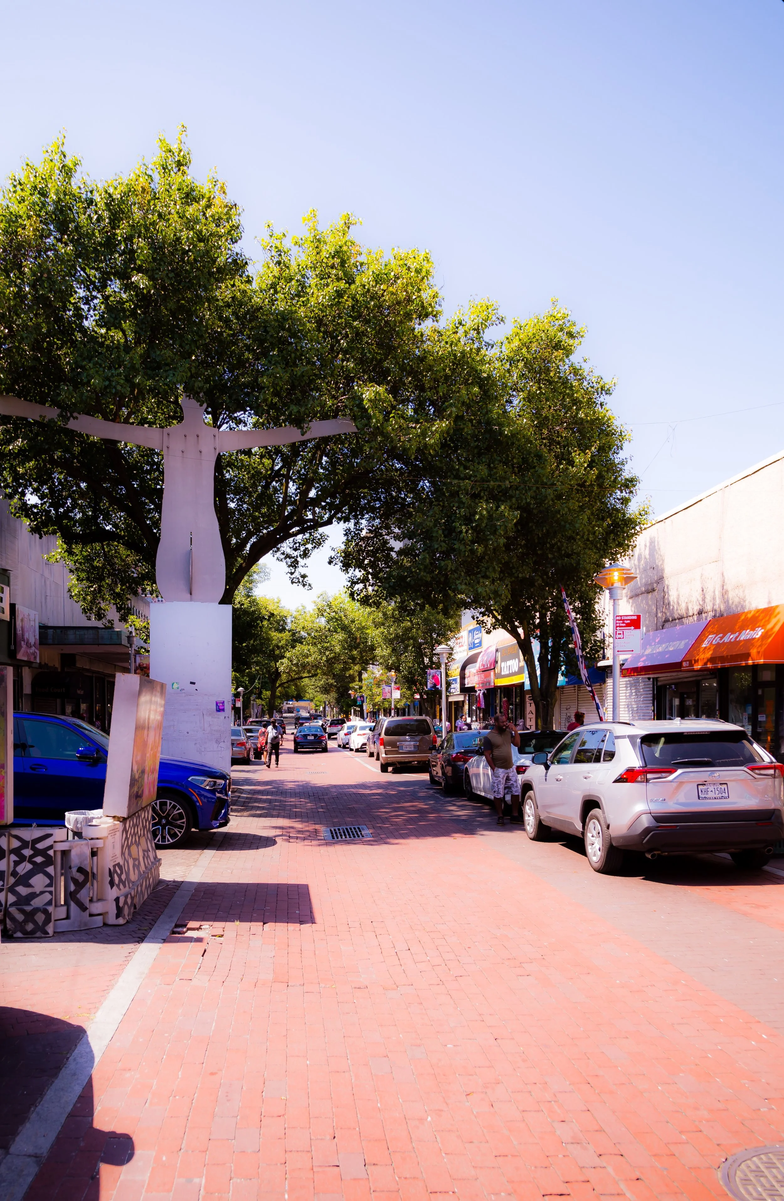 Street view with parked cars, trees, pedestrians, local shops, and a large sculpture of a human figure with outstretched arms.