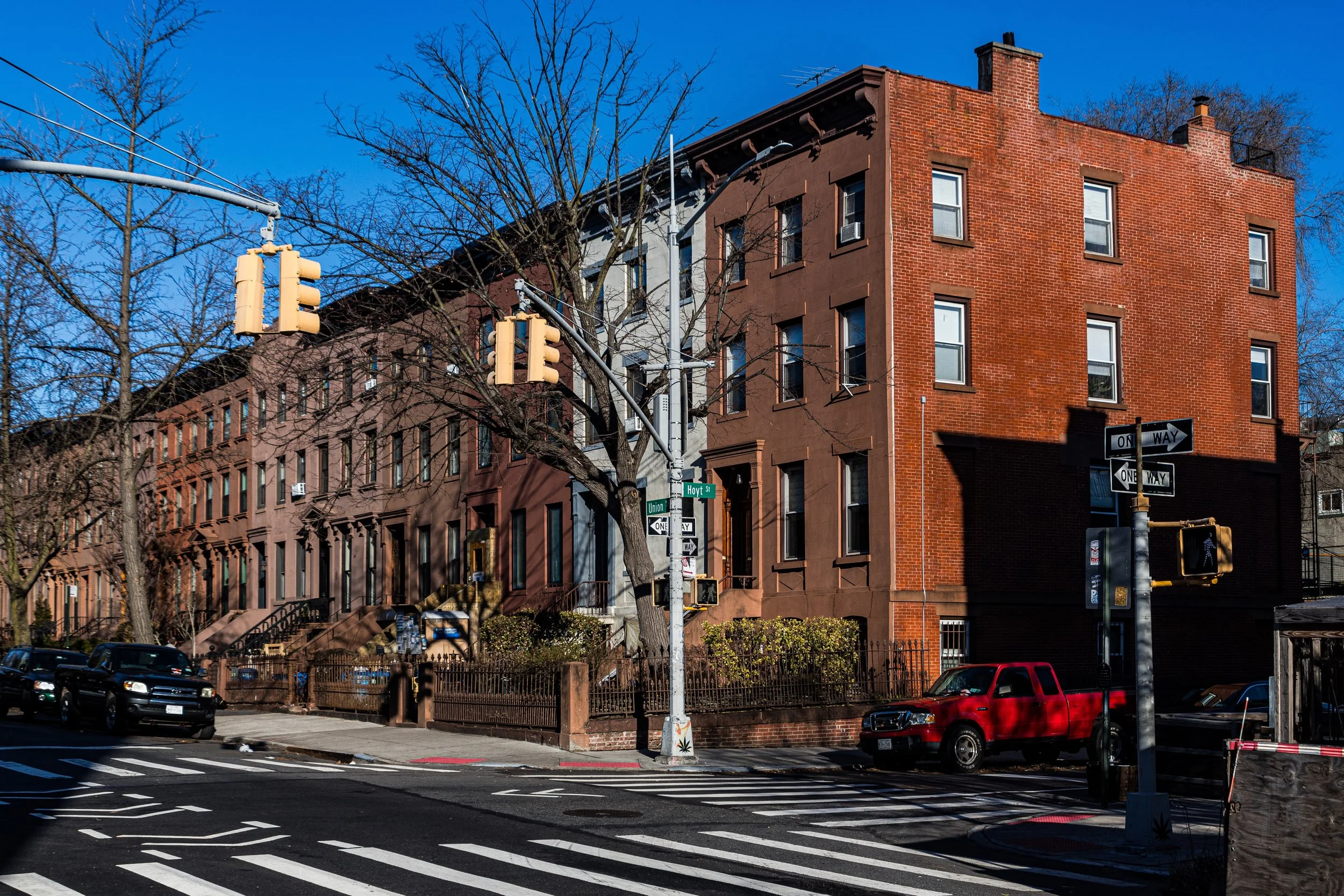 A street corner with multi-story brick buildings, traffic lights, street signs, and parked cars under a clear blue sky.