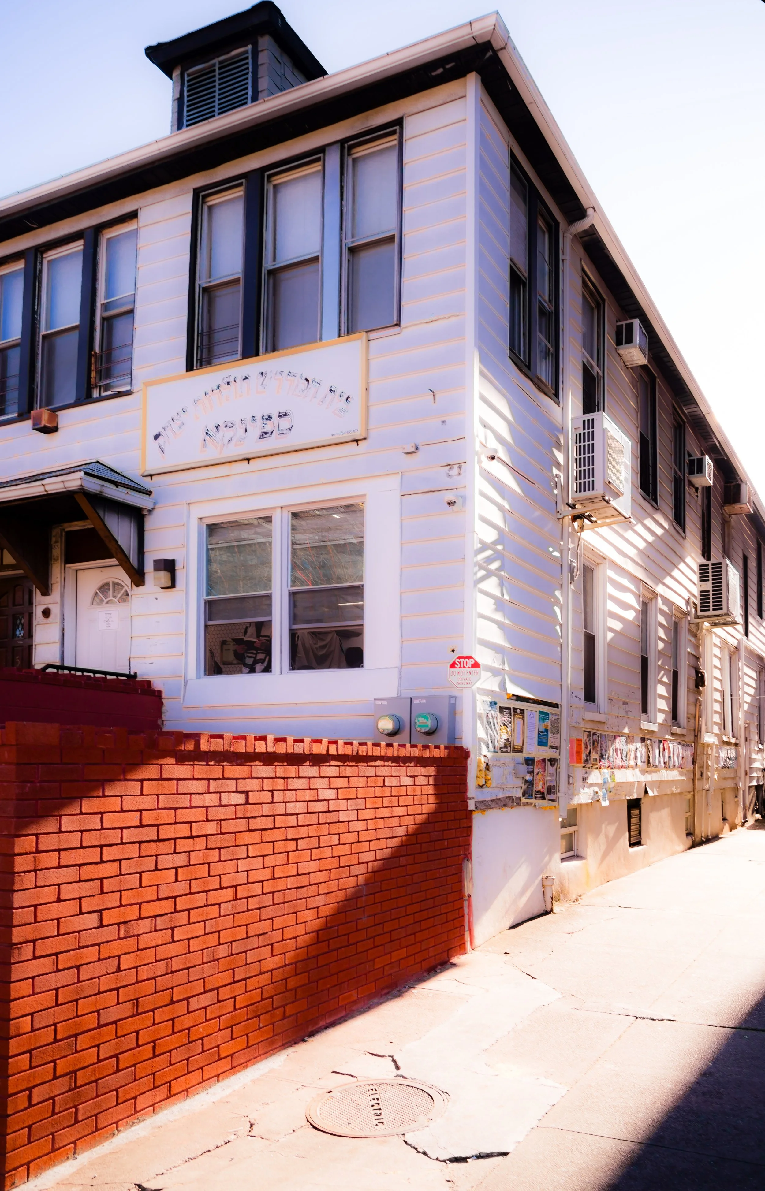 White two-story building with black window trims and air conditioning units, a brick wall in front, and a sidewalk beside it.