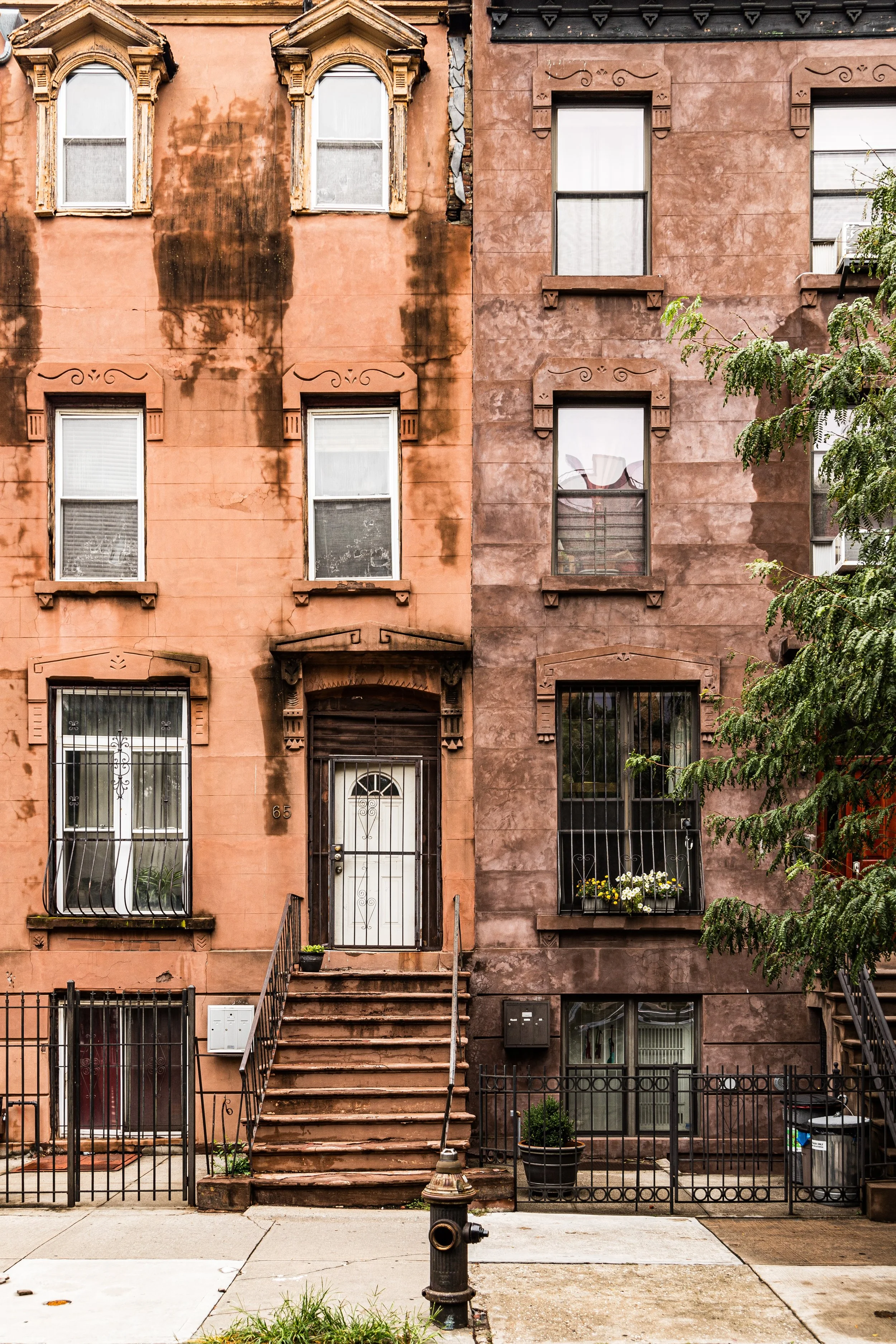 Old brick apartment building with three floors and stairs leading to the front door, featuring a fire hydrant and small plants on the sidewalk.