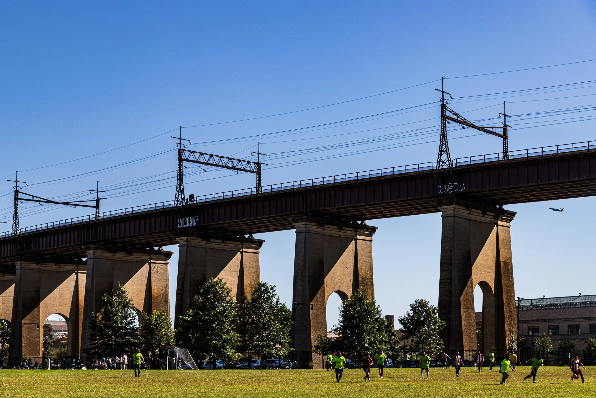 A sports field with players playing soccer, under a large bridge with multiple arches and electrical towers. There are trees and a few buildings in the background, and a plane flying in the sky.