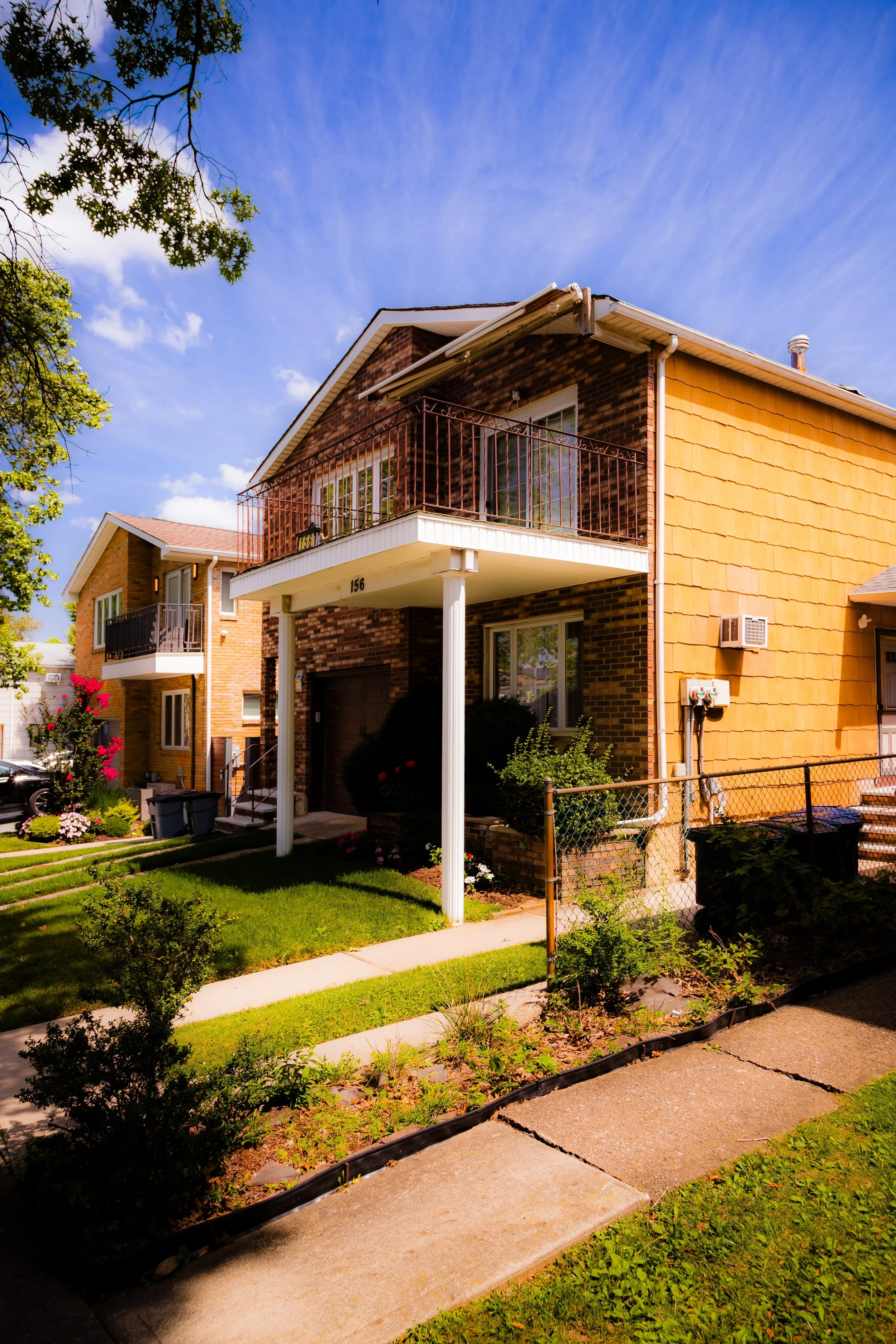 A residential two-story brick house with a small front porch and balcony, landscaped yard, sidewalk, and neighboring similar houses under a blue sky with some clouds.
