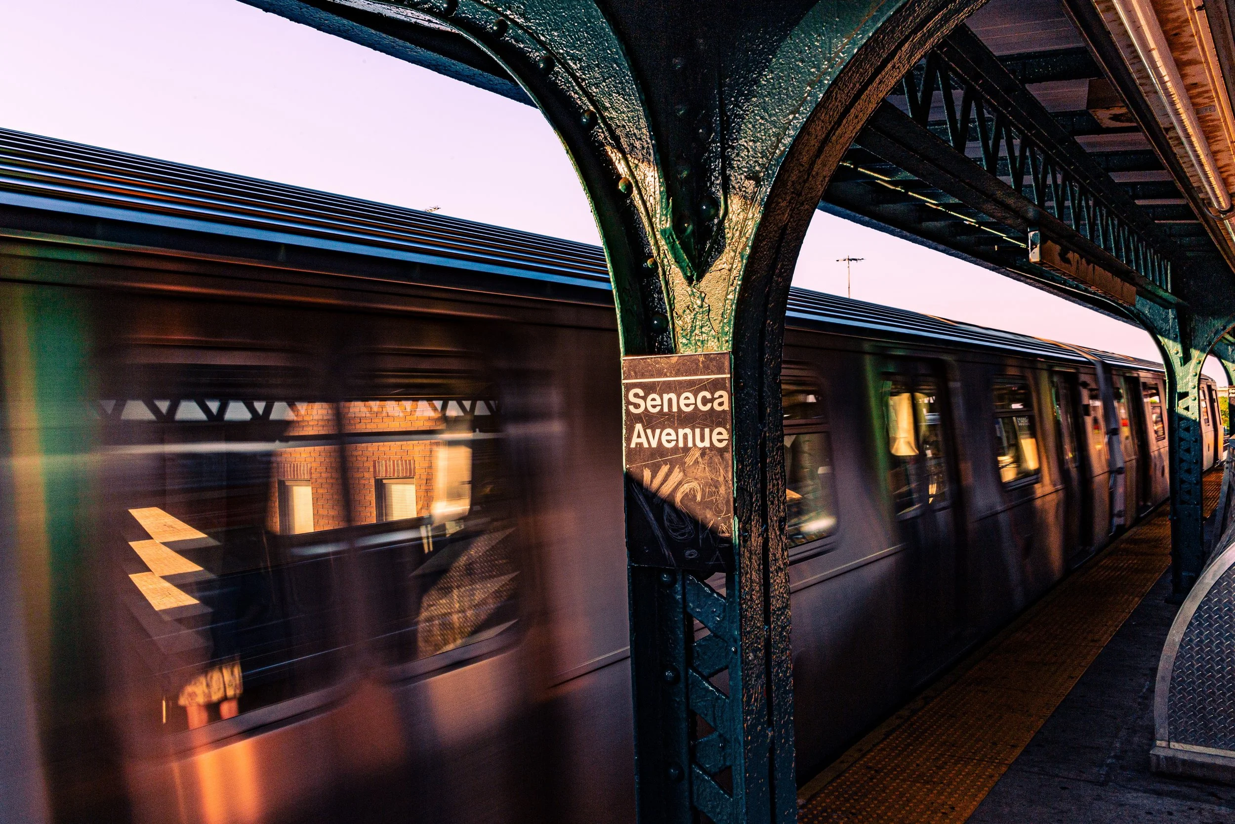 A moving subway train at Seneca Avenue station in New York, with the platform and a sign visible in the image during dusk.