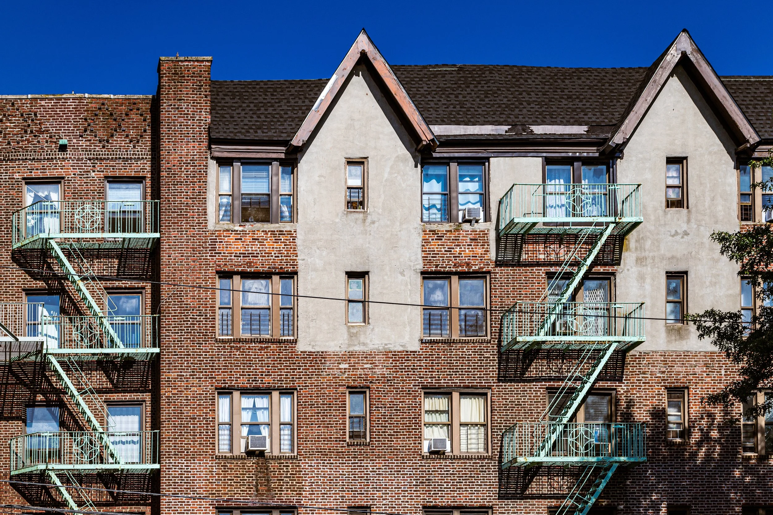 A red brick apartment building with fire escape stairs and multiple windows, some with air conditioning units, under a blue sky.