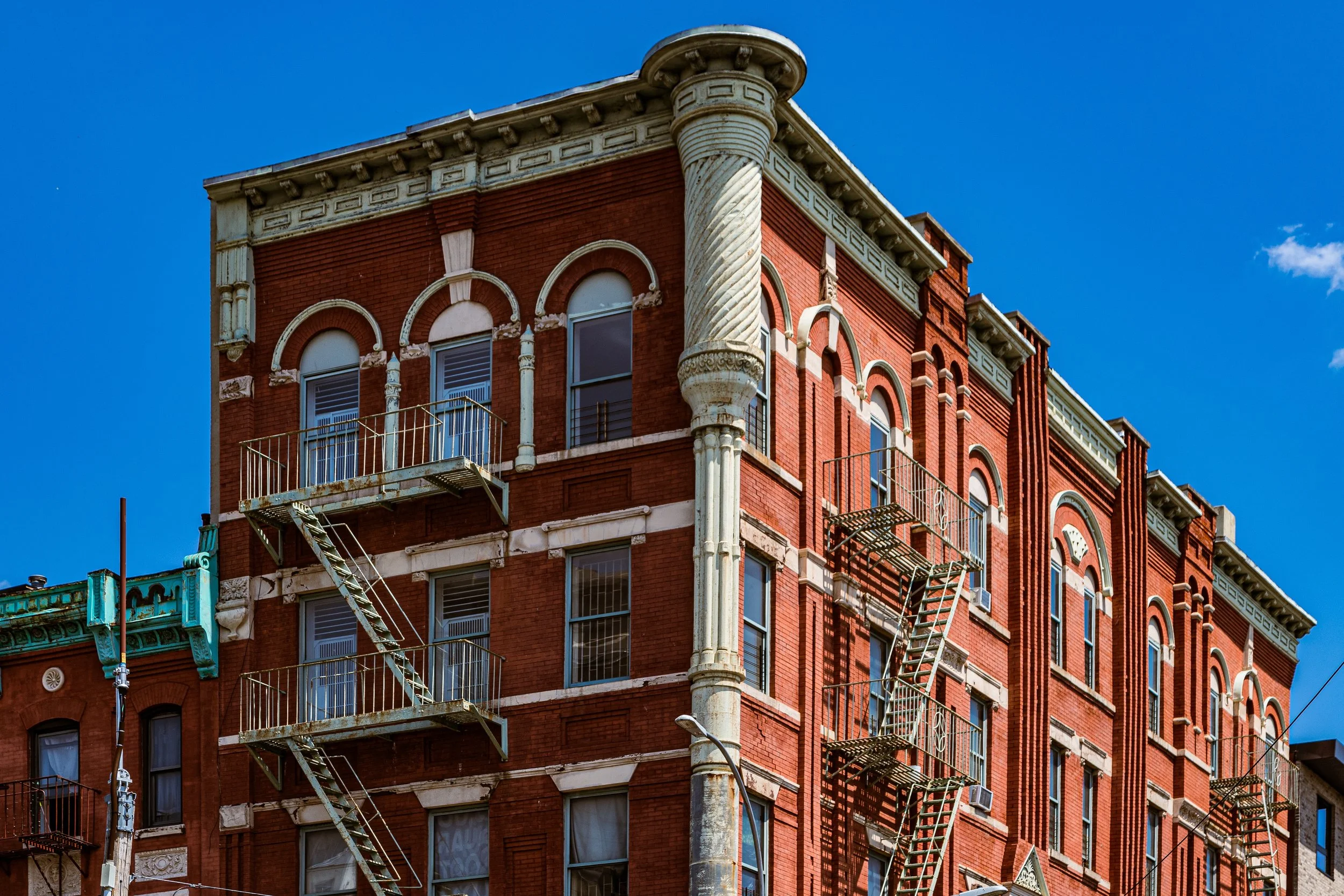 Red brick building with ornate architectural details and fire escape stairs on the exterior, against a bright blue sky.