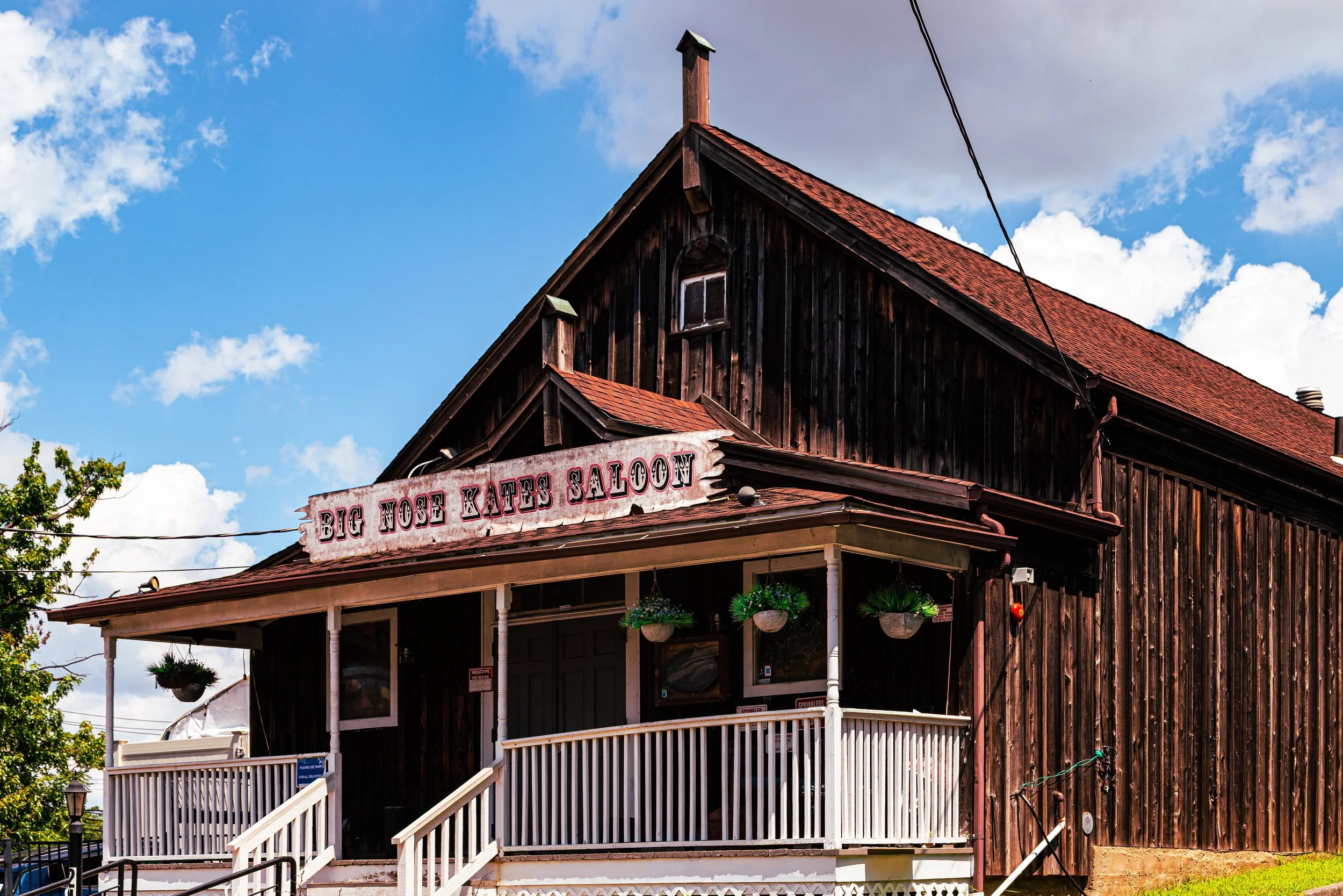 Old wooden building with a sign that reads 'Big Nose Kate's Saloon', white porch railing, hanging plants, under a partly cloudy sky.