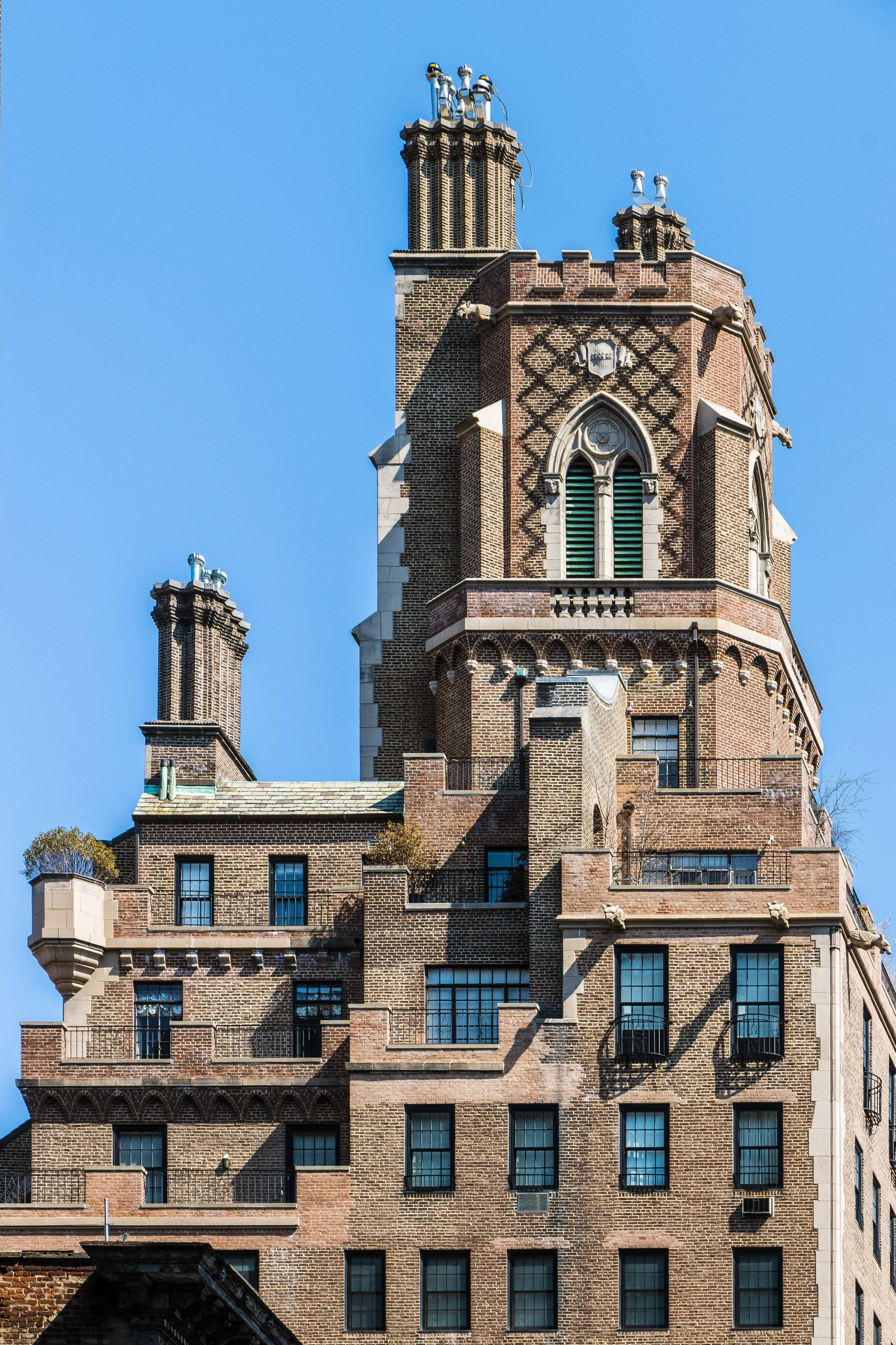 The image shows a large, ornate brick building with a prominent clock tower and multiple chimneys against a clear blue sky.