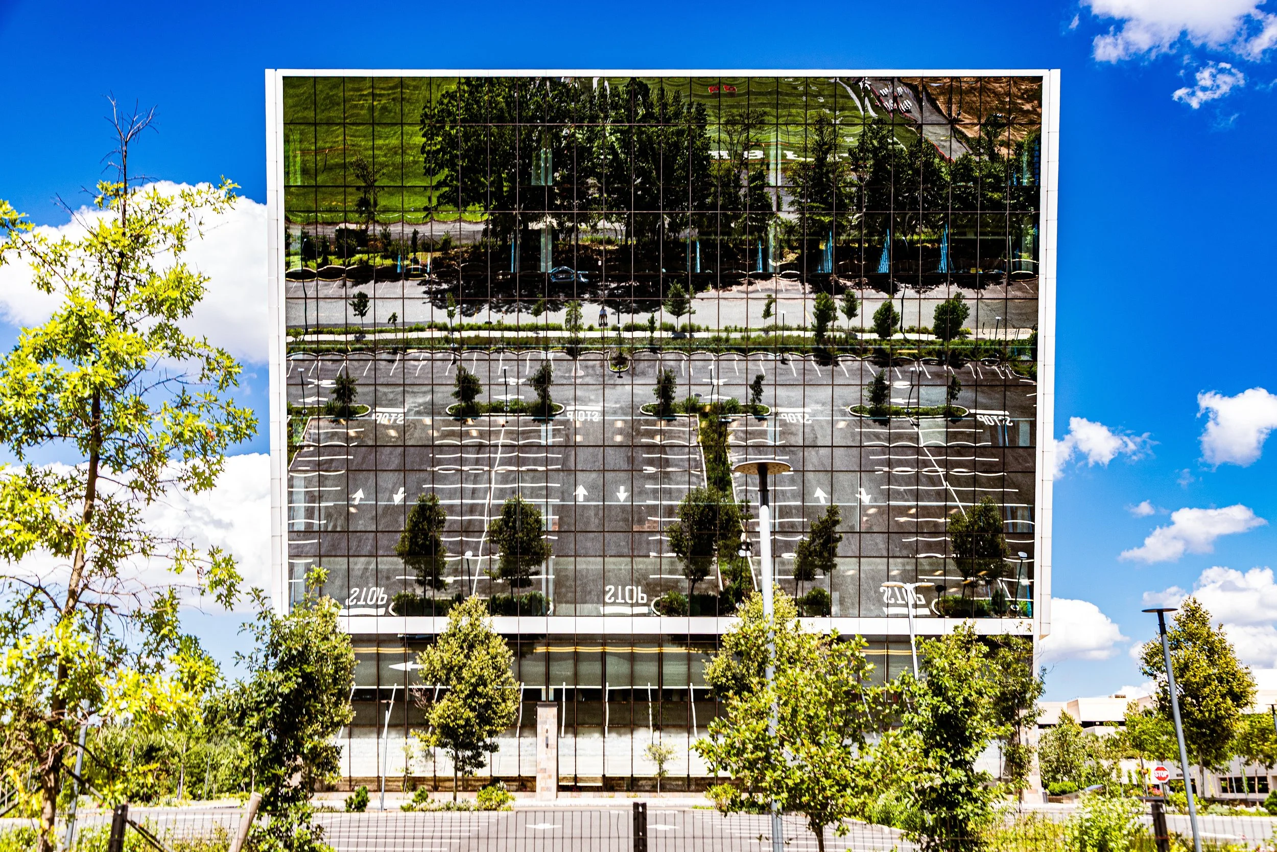 Reflective glass building mirrors the parking lot and trees in front of it on a bright, sunny day with a blue sky and white clouds.