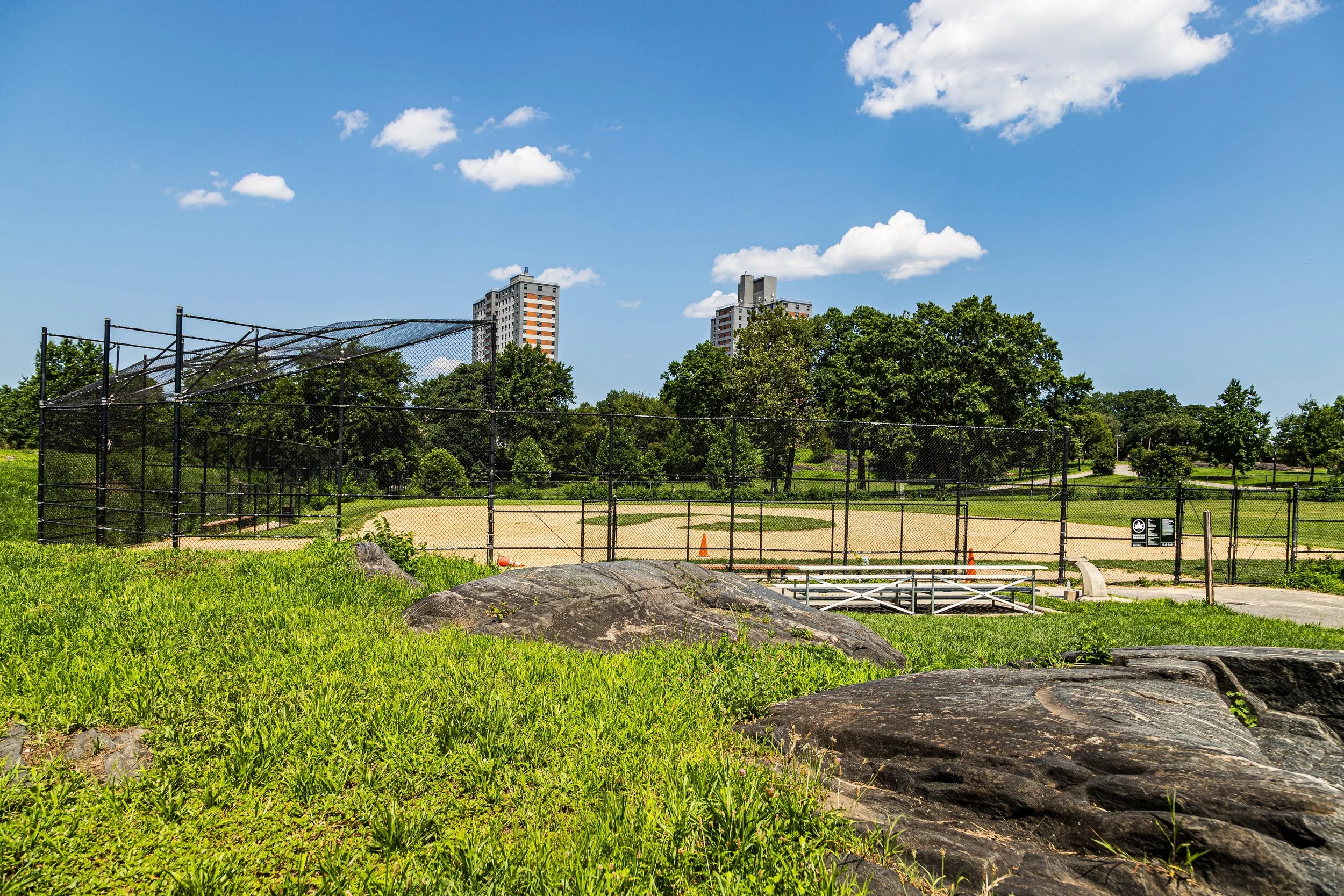 A baseball field with a fence, surrounded by green grass and trees, with two tall apartment buildings in the background under a blue sky with a few white clouds.