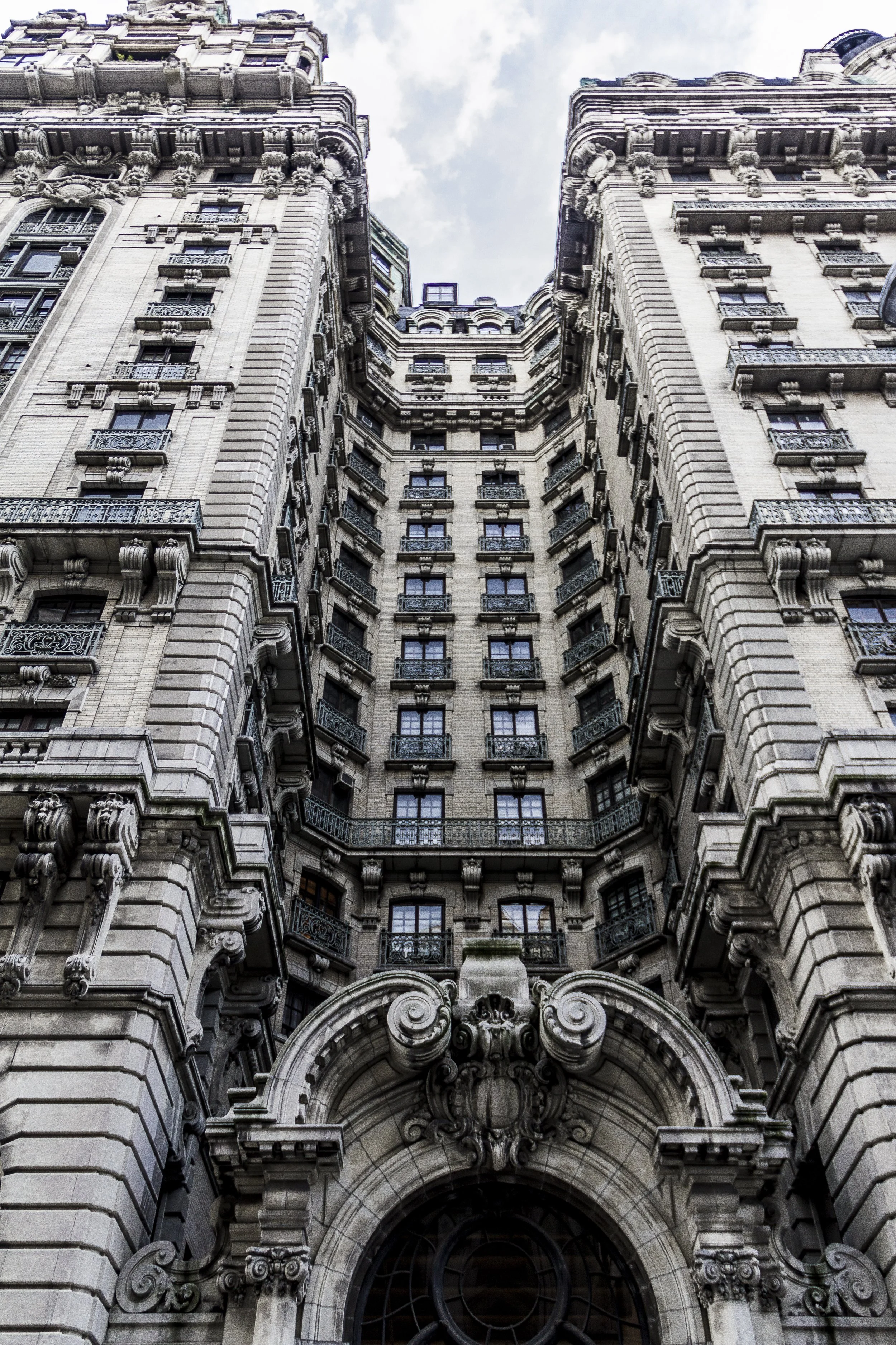 Looking up at a historic, ornate building with classical architecture, featuring intricate stone carvings, decorative pillars, balconies, and multiple windows, set against a partly cloudy sky.