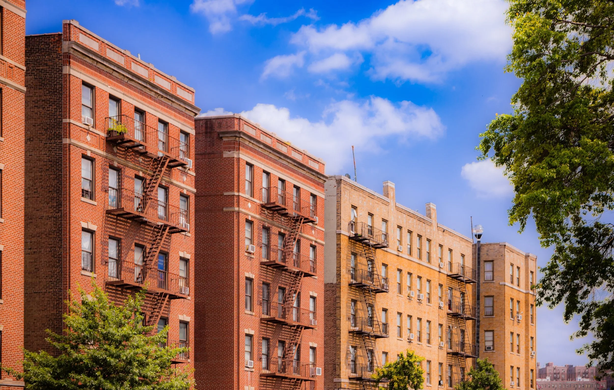 Multi-story brick apartment buildings with fire escapes on a sunny day, green trees in the foreground, and a blue sky with clouds.