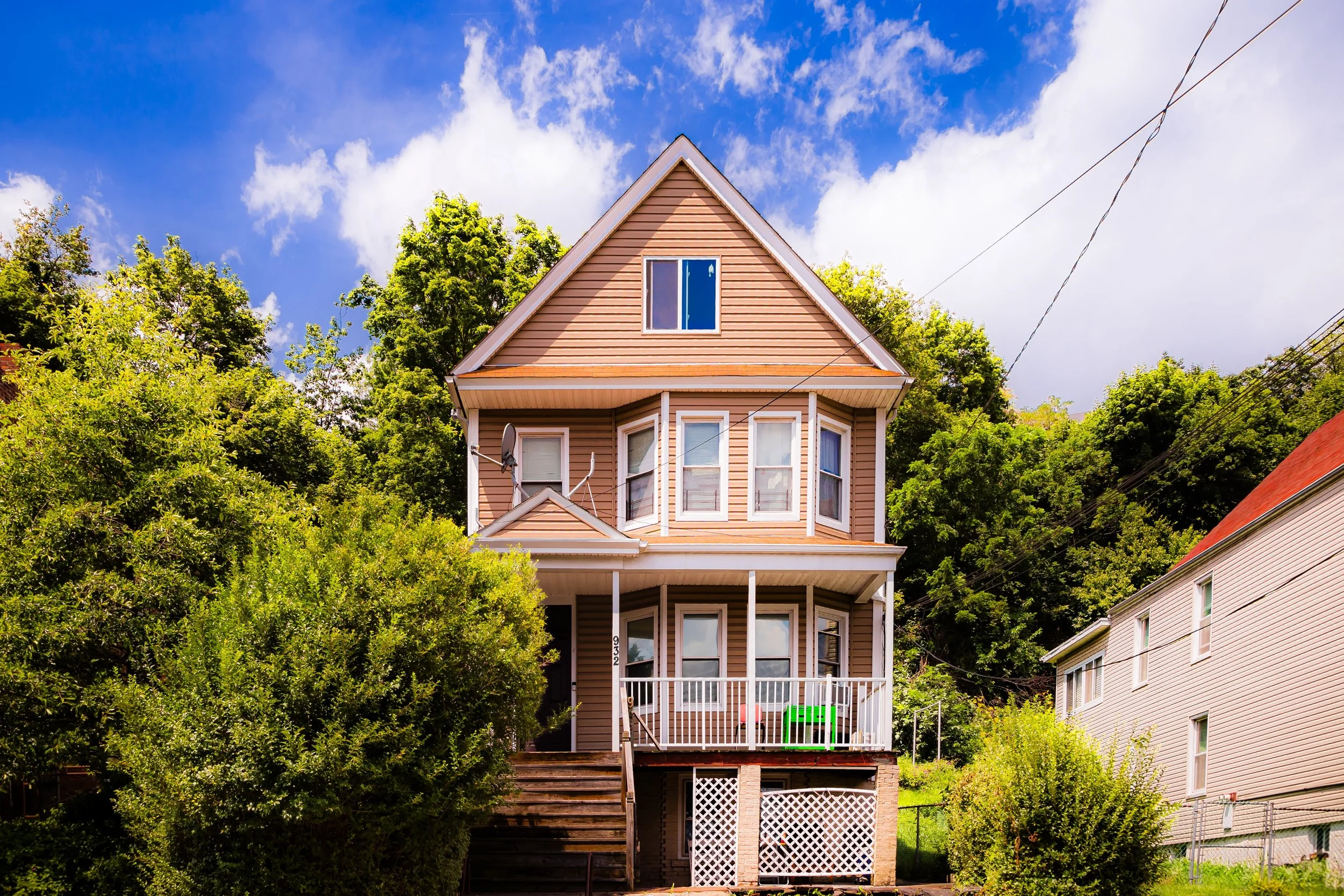 A three-story house with beige siding, a covered front porch, and trees in the background under a blue sky with clouds.