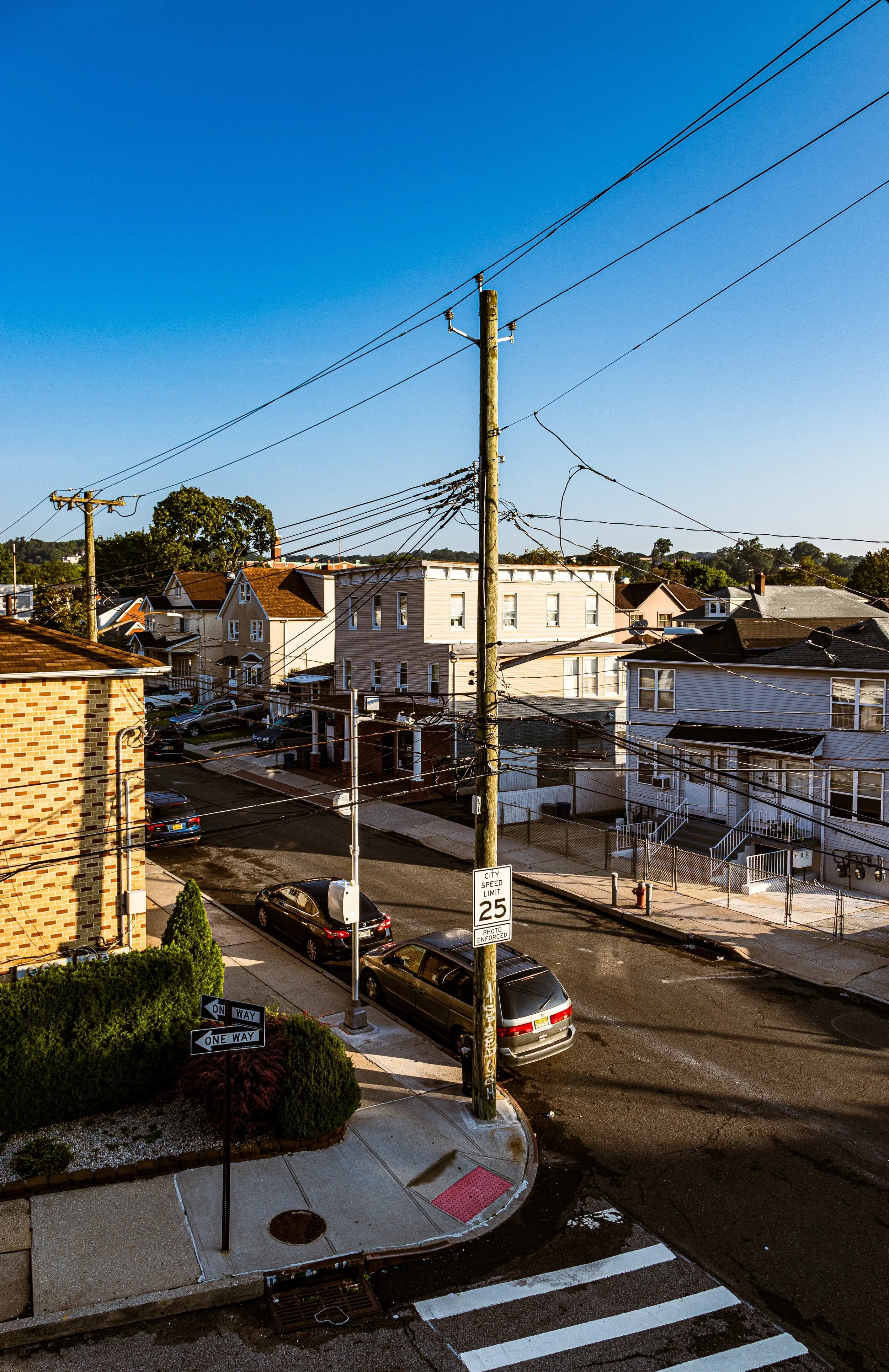 A street scene in a residential neighborhood with parked cars, utility poles with wires, and houses under a clear blue sky.