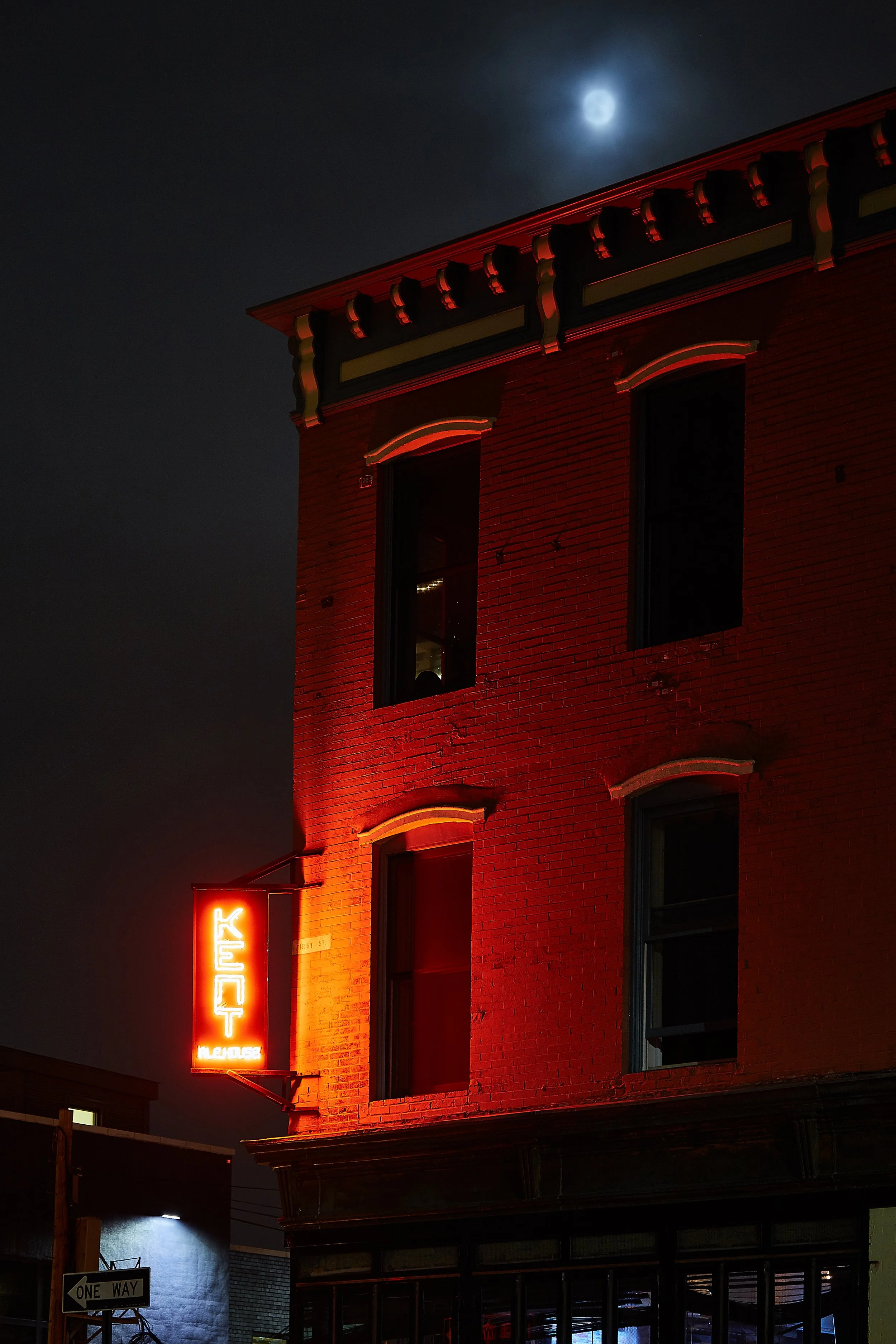 Nighttime scene of a red brick building with a neon sign that reads 'KENT' in all caps and 'HEADHOUSE' below it. The building has several windows, some with lights on. The moon is visible in the cloudy night sky.