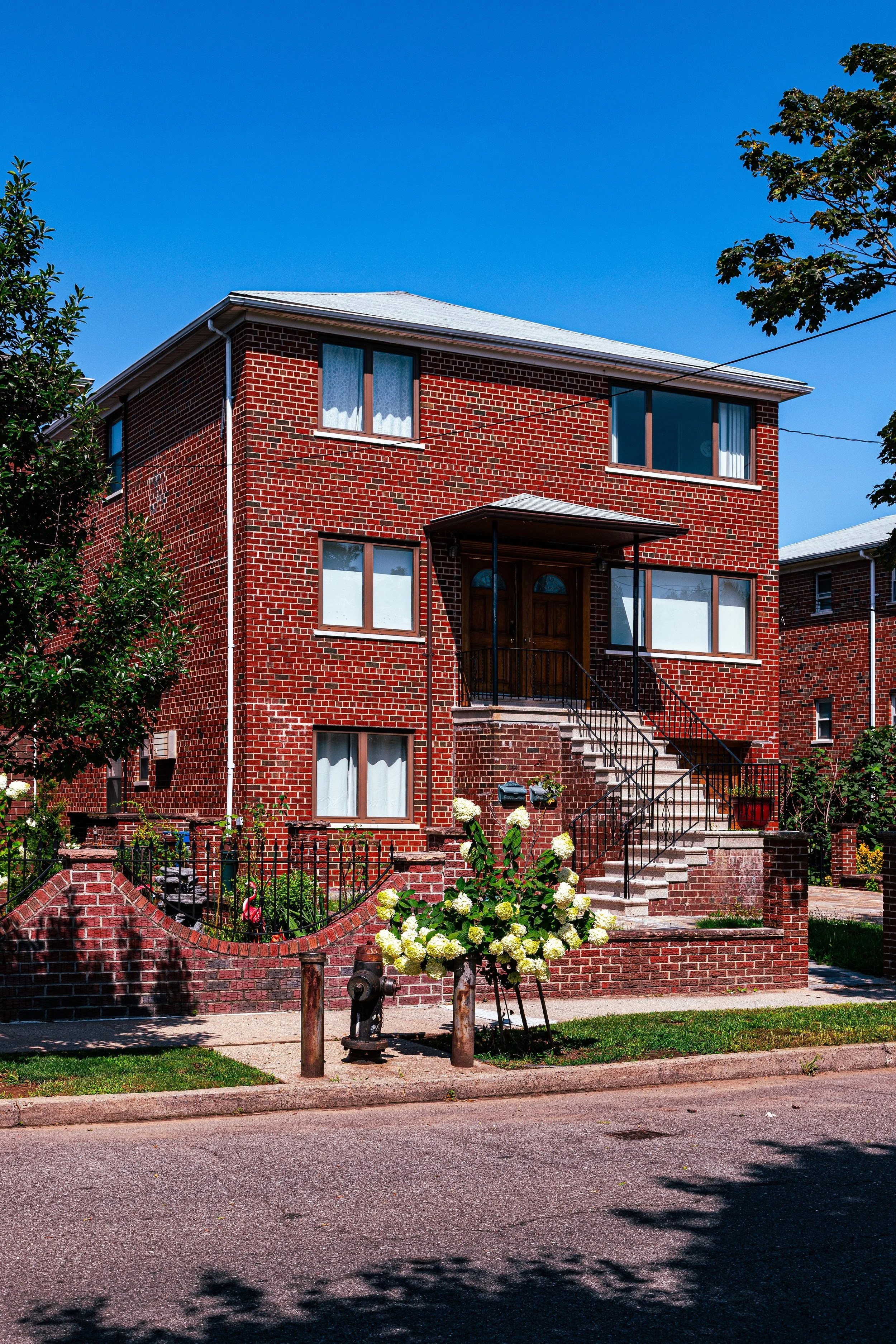 A three-story brick house with stairs leading to the front door, surrounded by trees and flowers, under a bright blue sky.