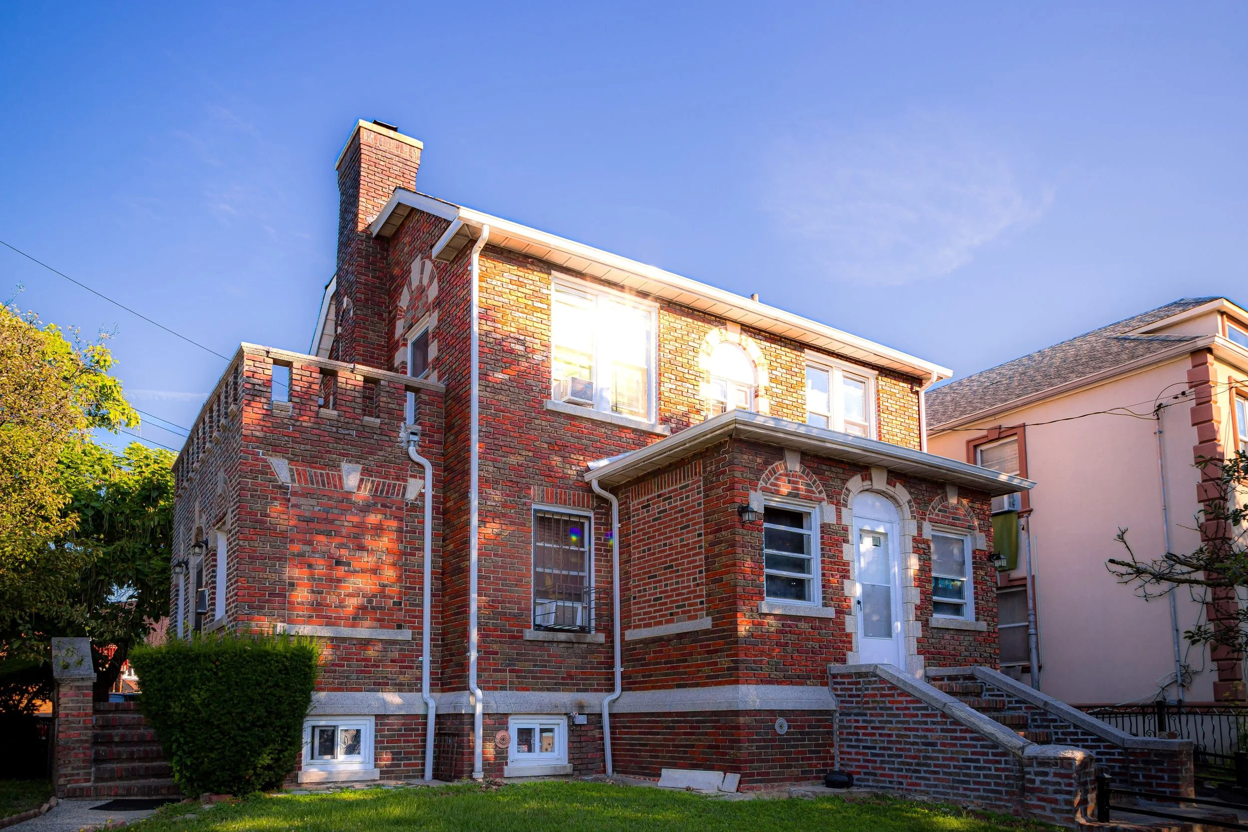 A brick house with a chimney, multiple windows, a white front door, and a small front staircase, under a clear blue sky with sunlight reflecting off the windows.
