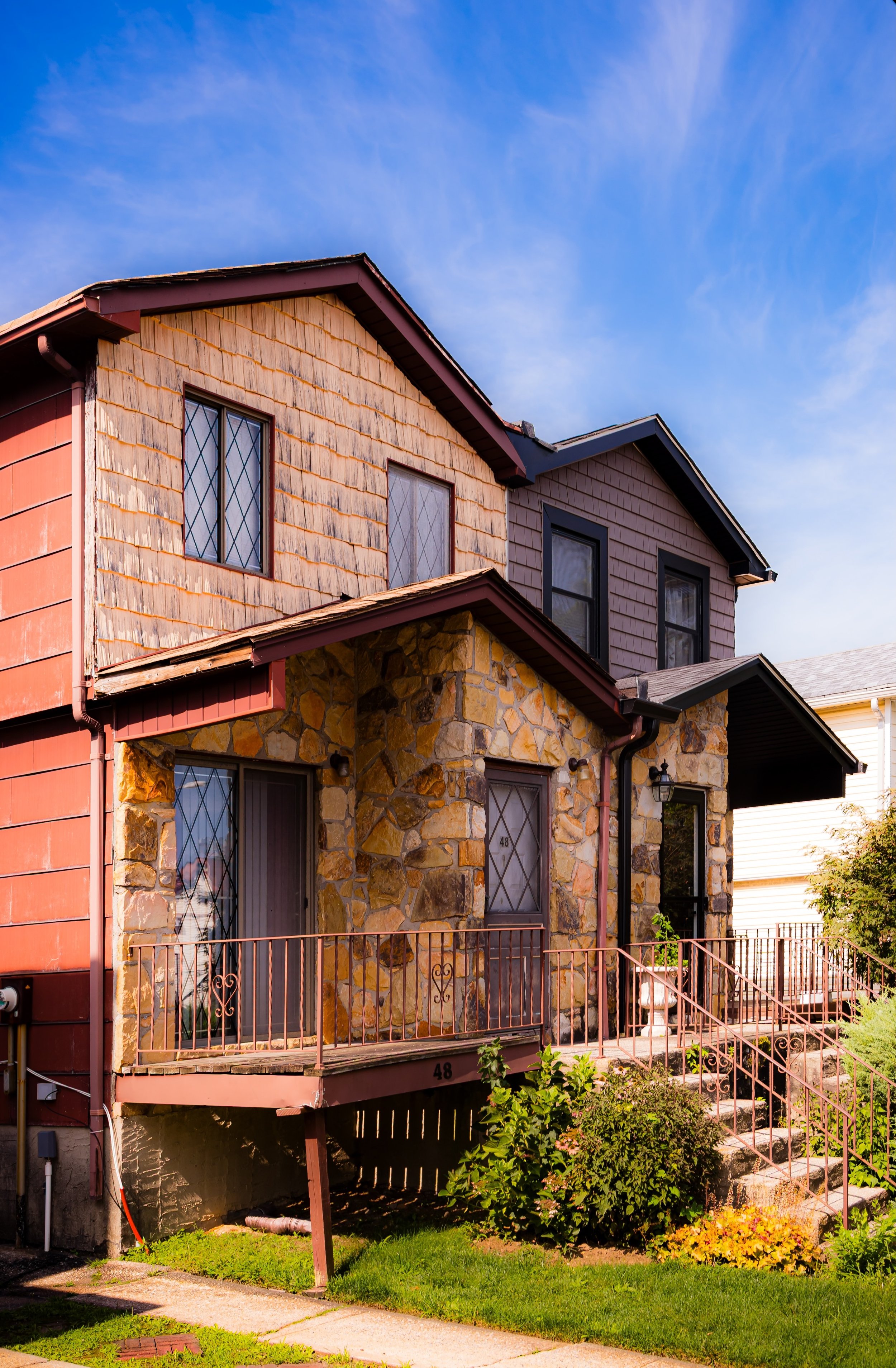 A multi-story house with a stone façade on the lower level and wooden siding on the upper levels, featuring windows with diamond-shaped panes, a small front porch with steps, a metal railing, and a lush garden with bushes and flowers, under a bright blue sky.
