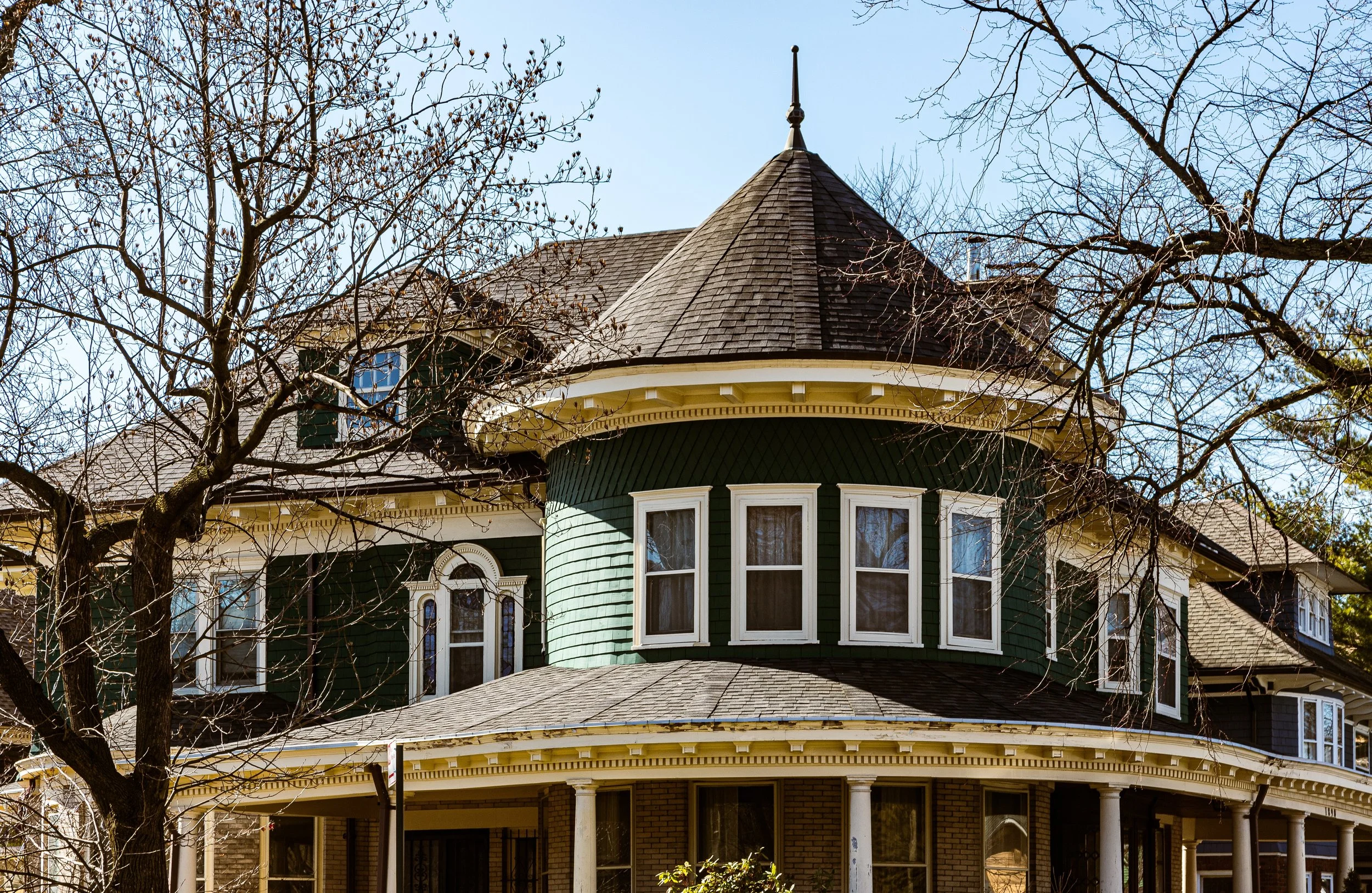 Victorian-style house with a domed turret, green siding, white trim, multiple windows, trees without leaves, and a clear blue sky.