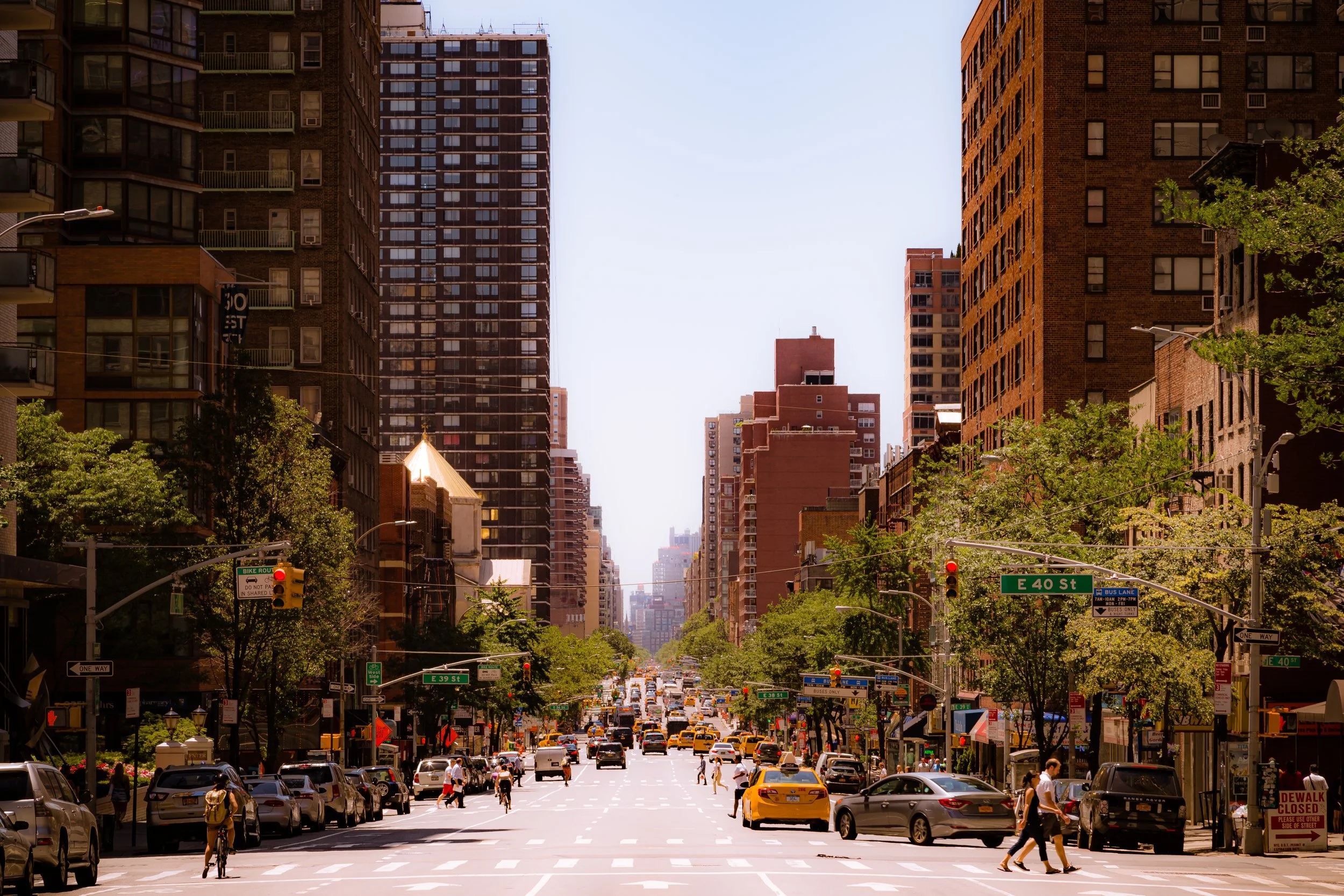 Busy city street with cars, yellow taxis, and pedestrians under tall buildings and green trees, indicating an urban setting on a sunny day.
