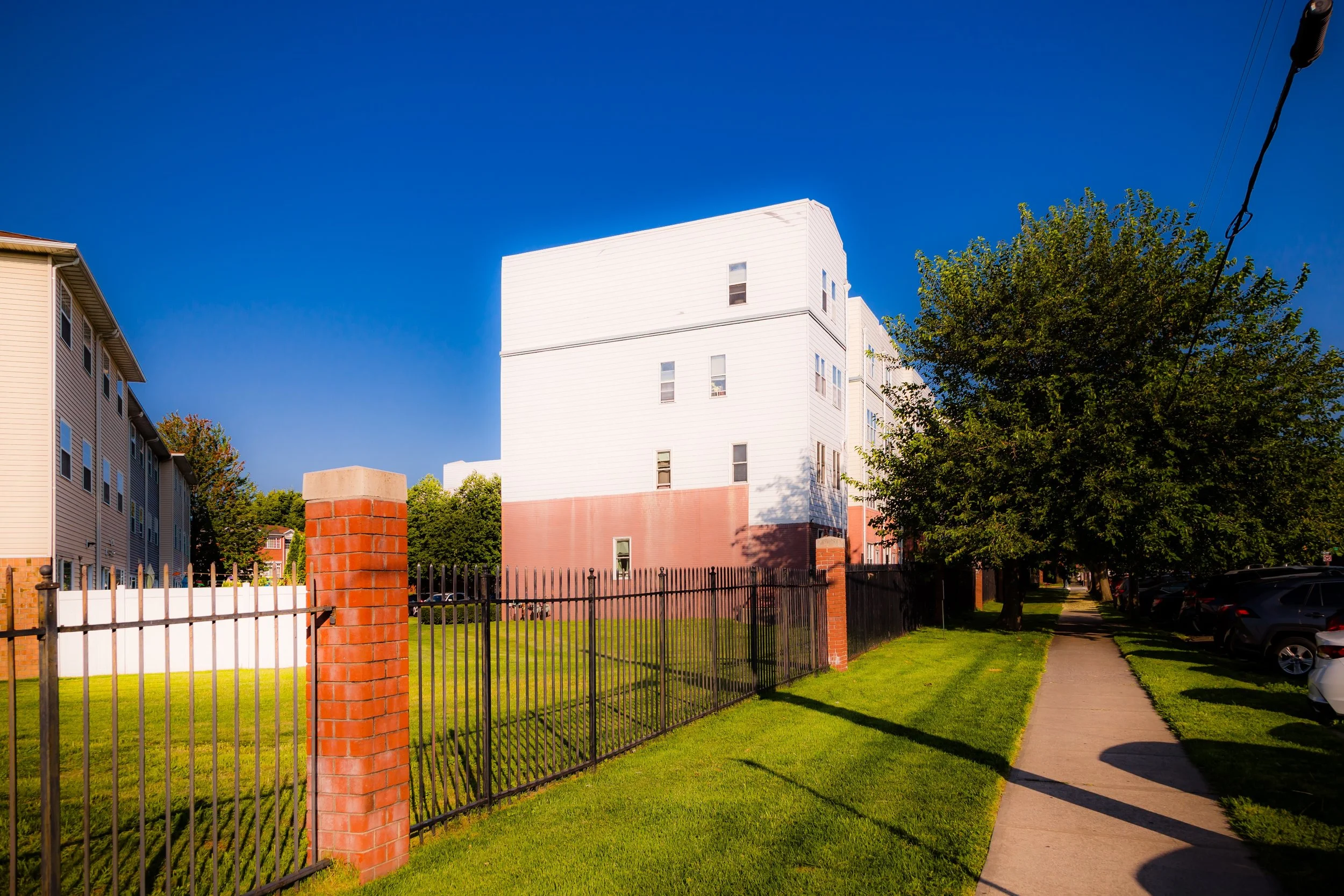 A sidewalk running alongside a grassy area bordered by a black metal fence, with a large house in the background and a tree on the right, under a clear blue sky.