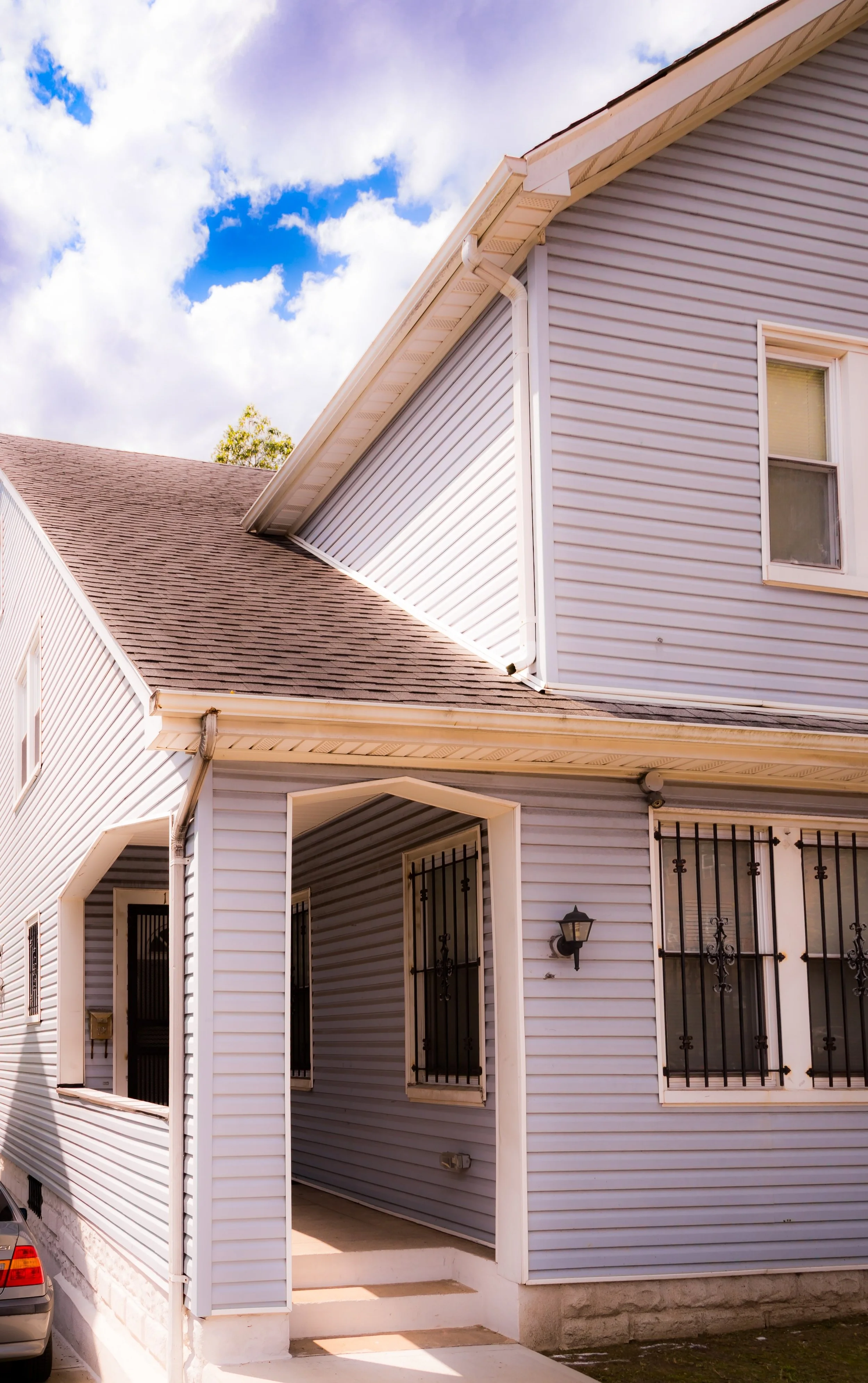 Front view of a light blue house with white trim, metal security bars on windows, a porch with steps, and a wall-mounted light fixture, under a partly cloudy sky.