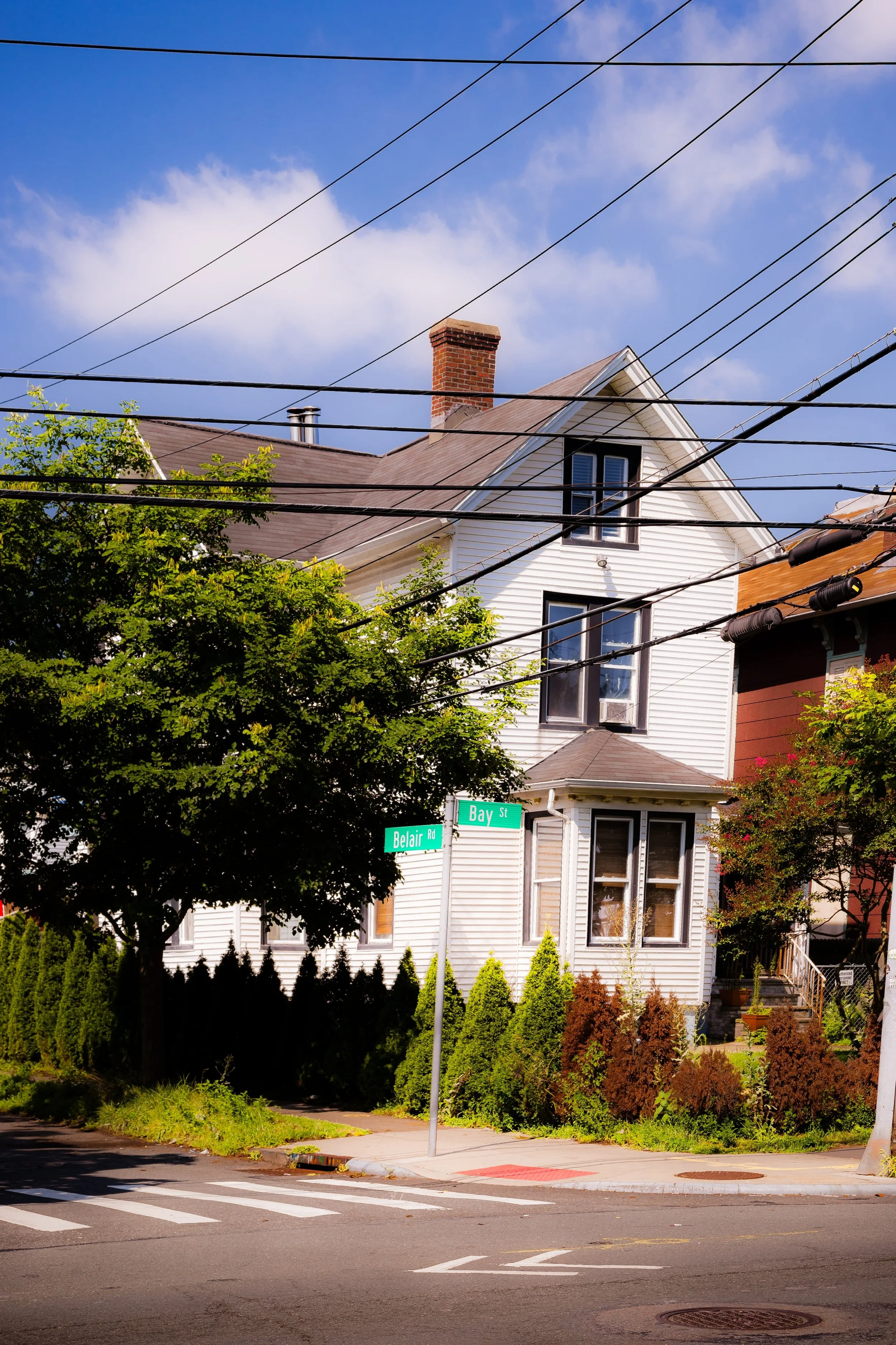 A white, three-story house on the corner of Belair Road and Bay Street, with power lines overhead, trees, and a corner sidewalk with crosswalk markings.
