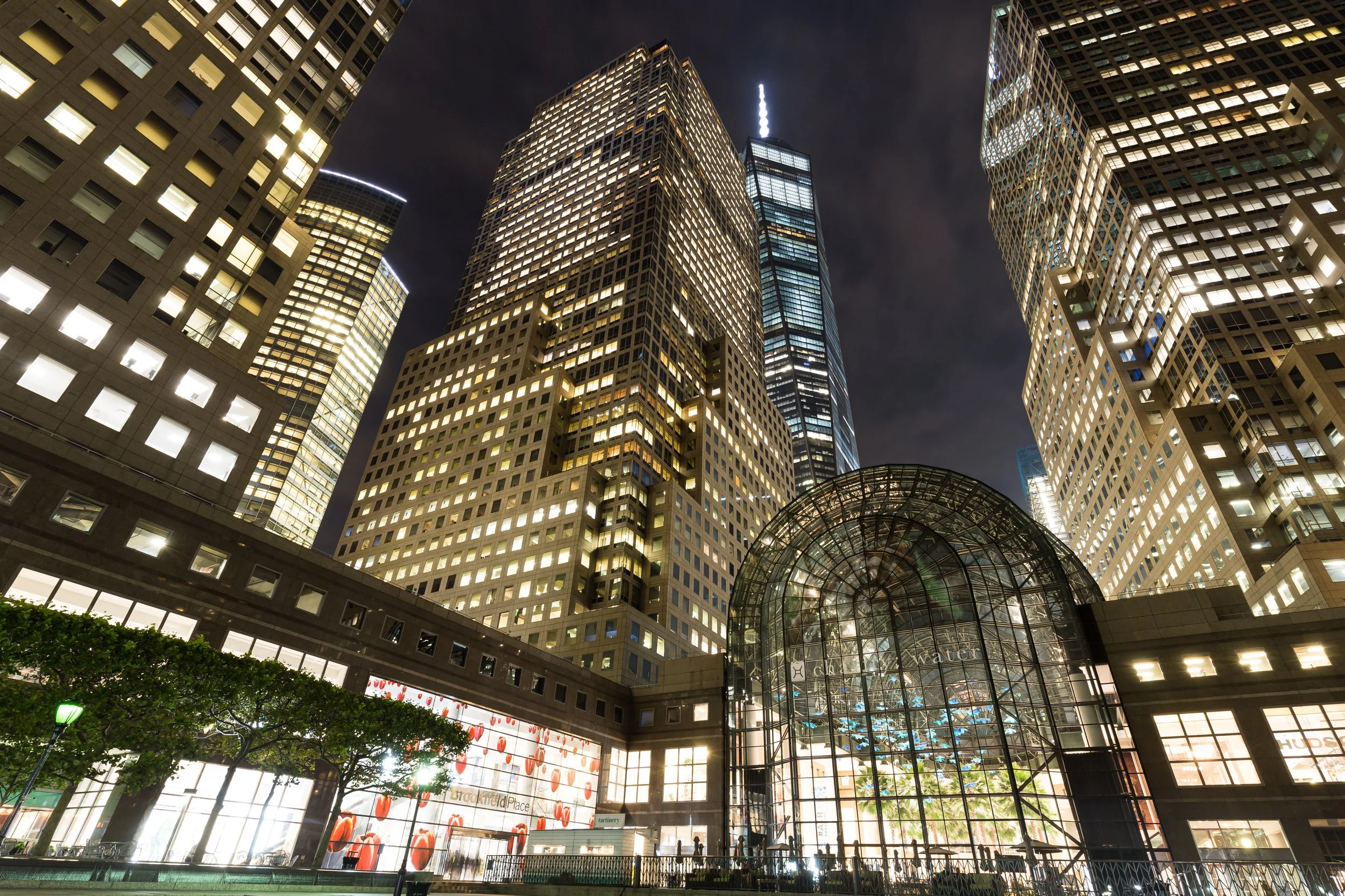 Night view of skyscrapers in a city, with illuminated windows and a glass entrance building at the bottom, in downtown Manhattan.