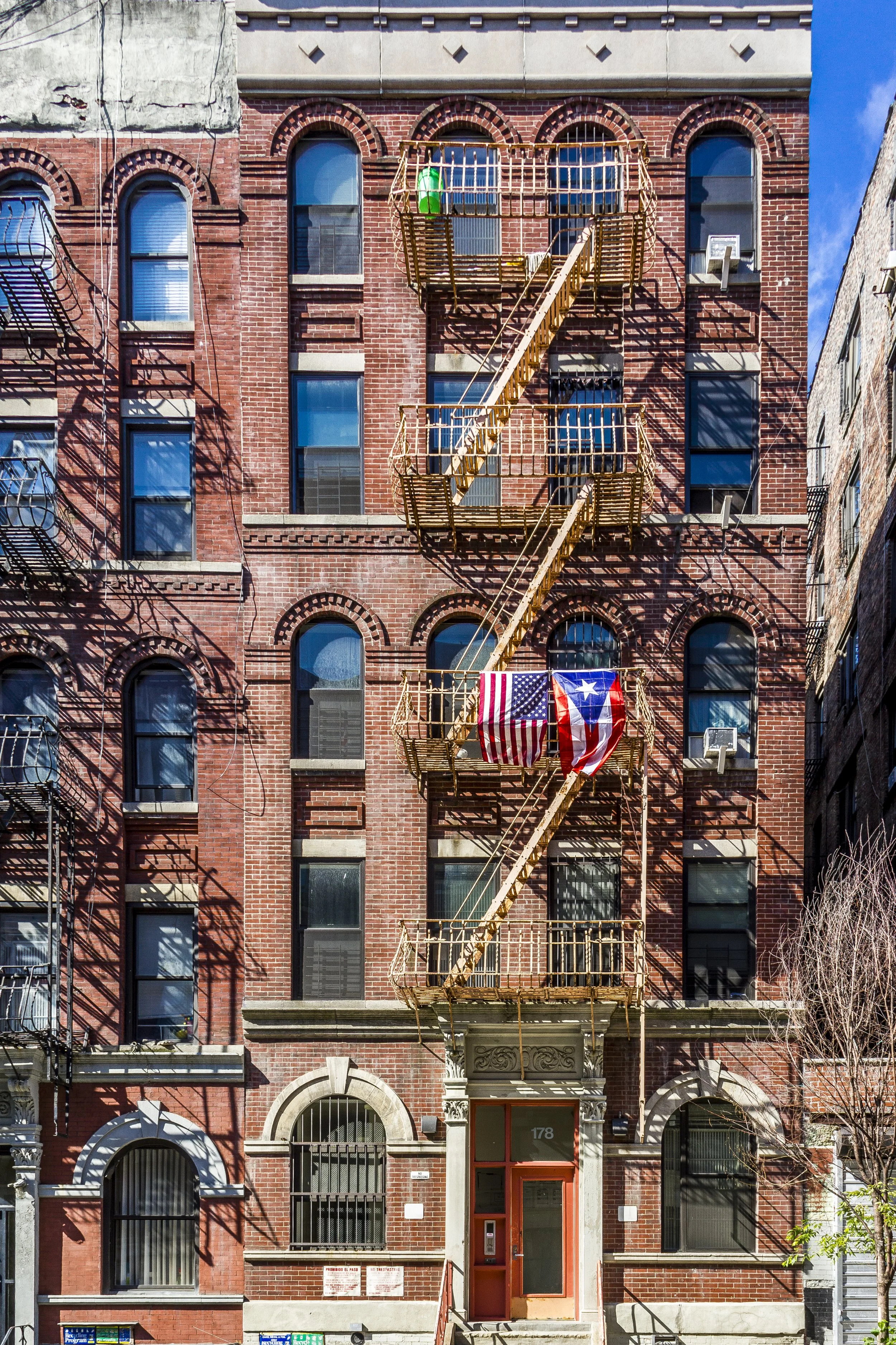 A red brick building with four floors, featuring arched windows with black bars and fire escape stairs with flags hanging from the middle landing. The building has ornate stonework around the entrance and fire escape, and shadows cast by fire escape embellishments.
