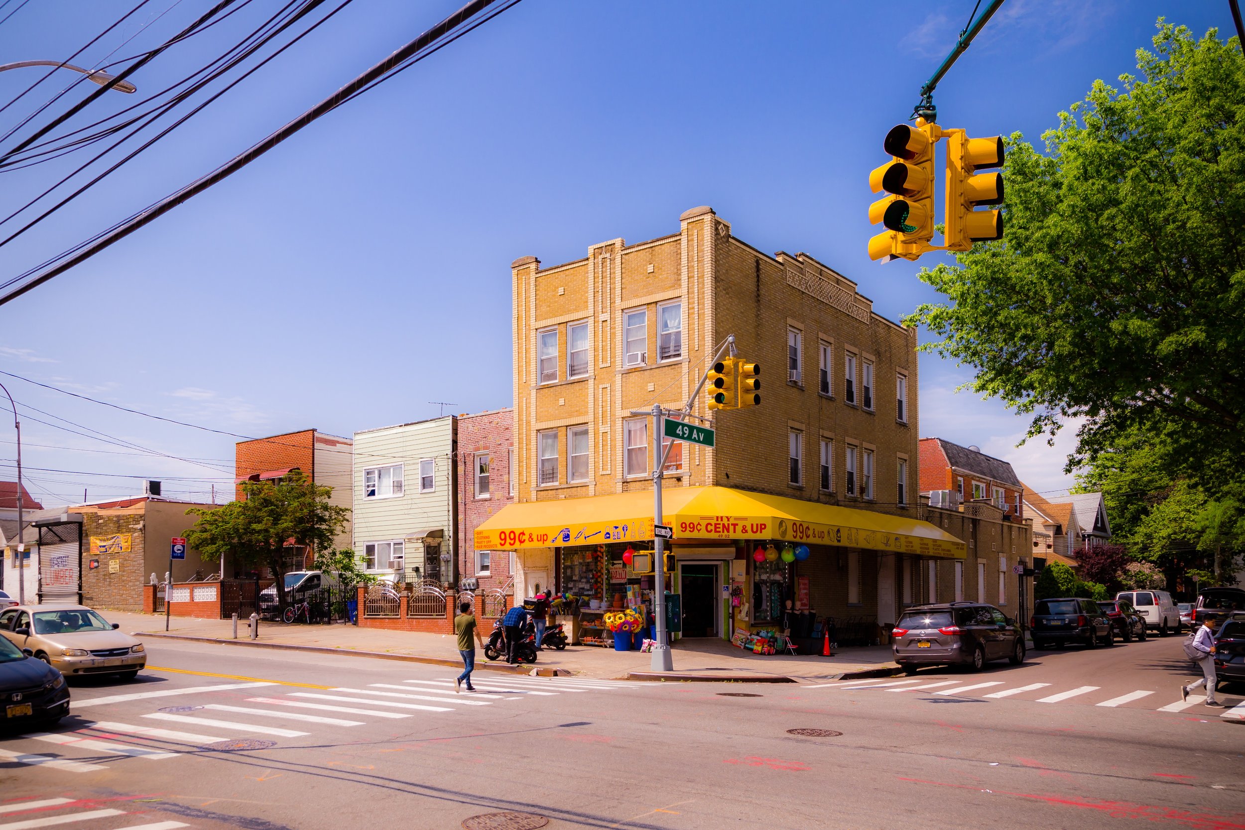 Corner building with a yellow awning displaying prices, at the intersection of 49th Avenue and a nearby street, with pedestrians crossing and cars parked and driving by.