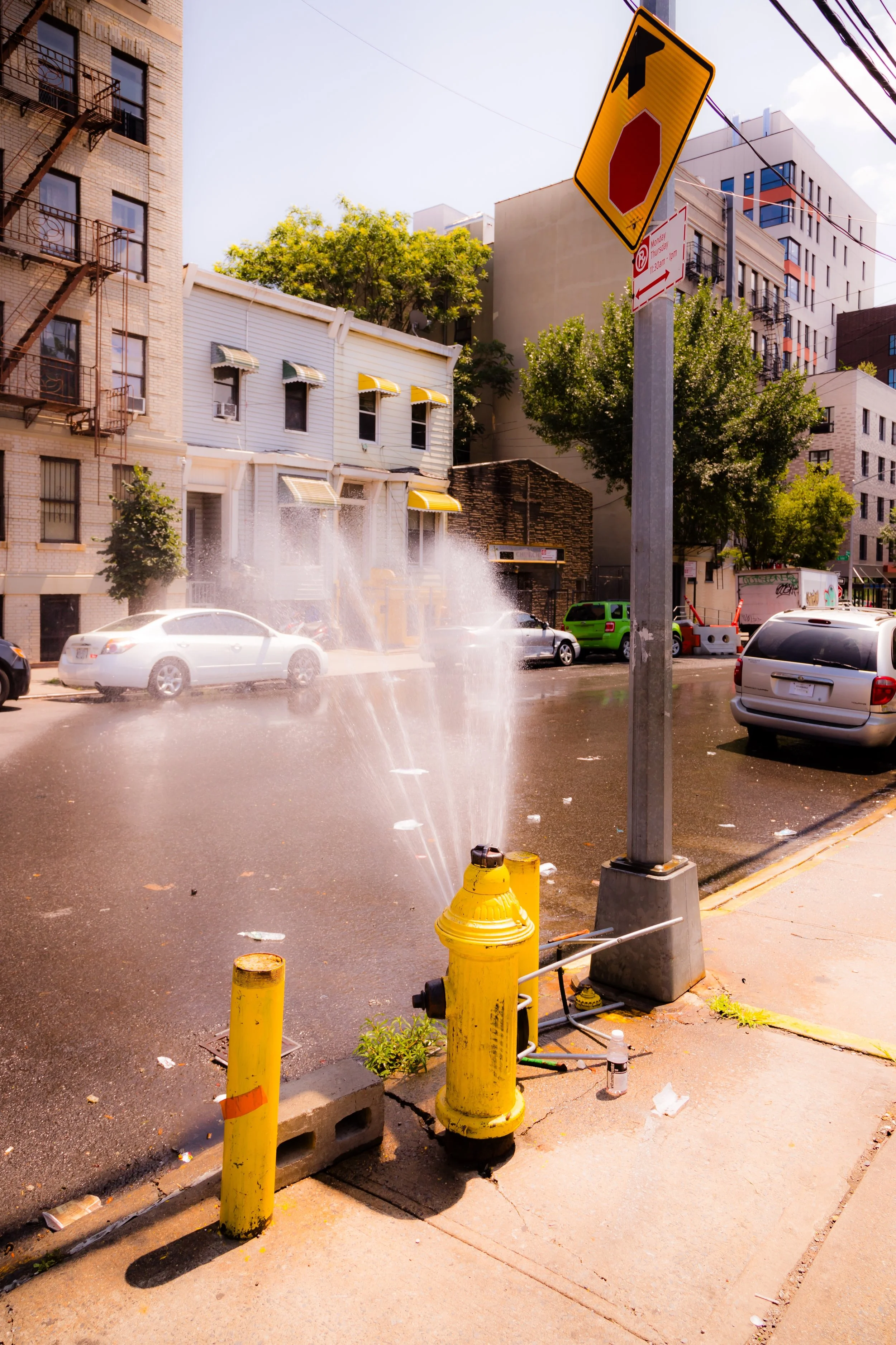 A yellow fire hydrant on a city street with water spraying out of it, parked cars, trees, and buildings in the background.