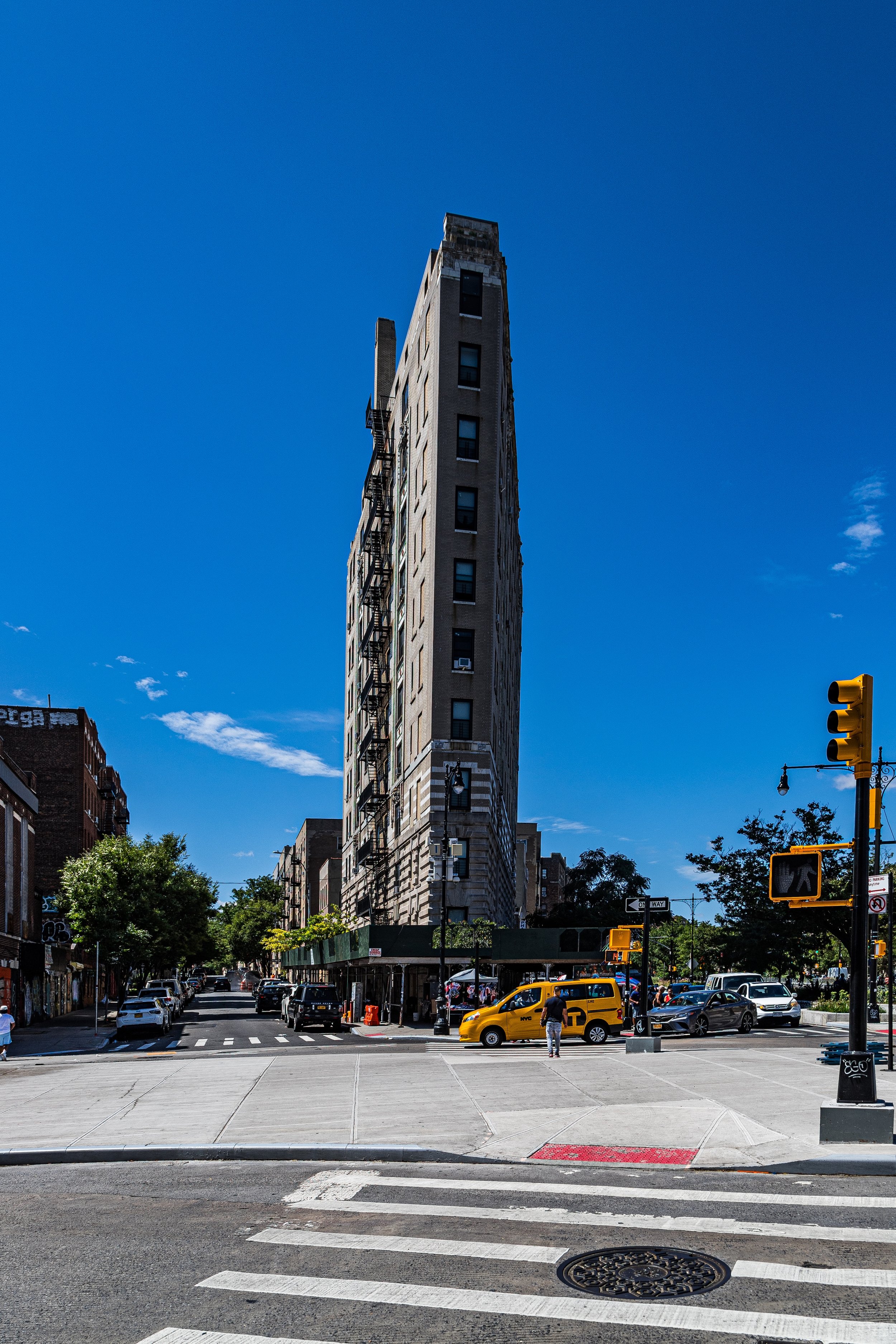 Tall, narrow building at the corner of a city street with cars and pedestrians, under a clear blue sky.