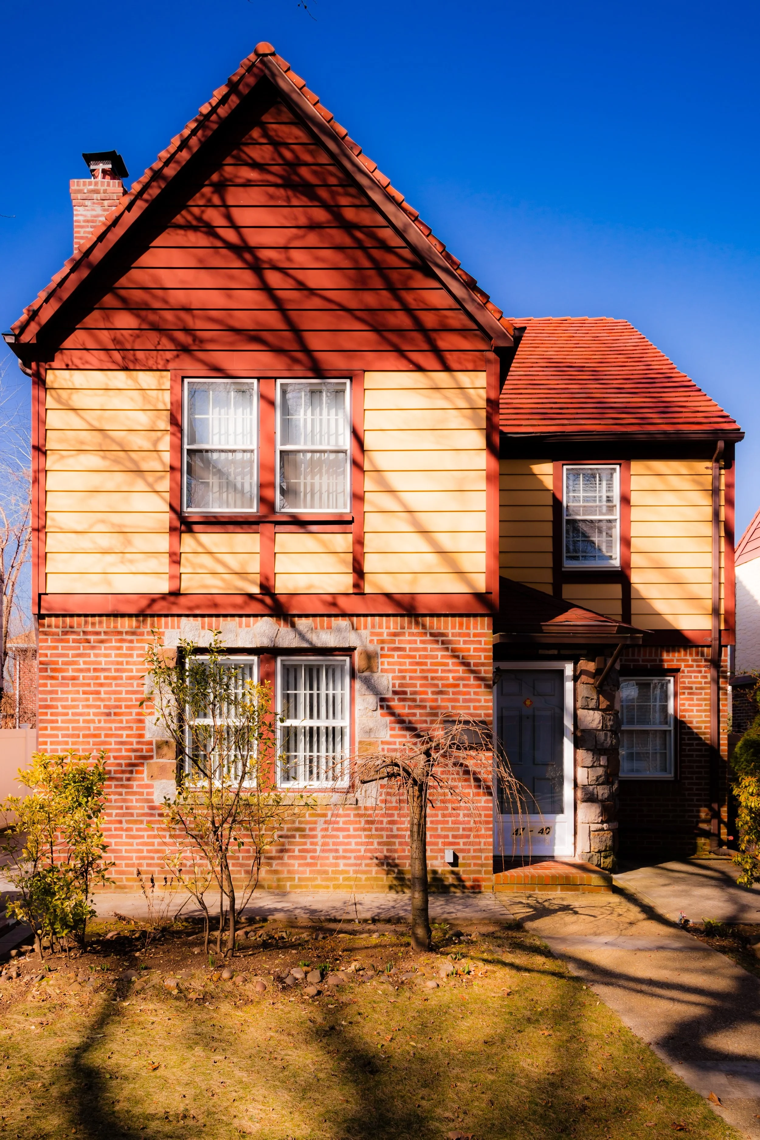 A two-story house with a brick ground floor and a yellow and red wooden upper floor, featuring four windows and a slate stone front door, situated in a suburban neighborhood under a clear blue sky.