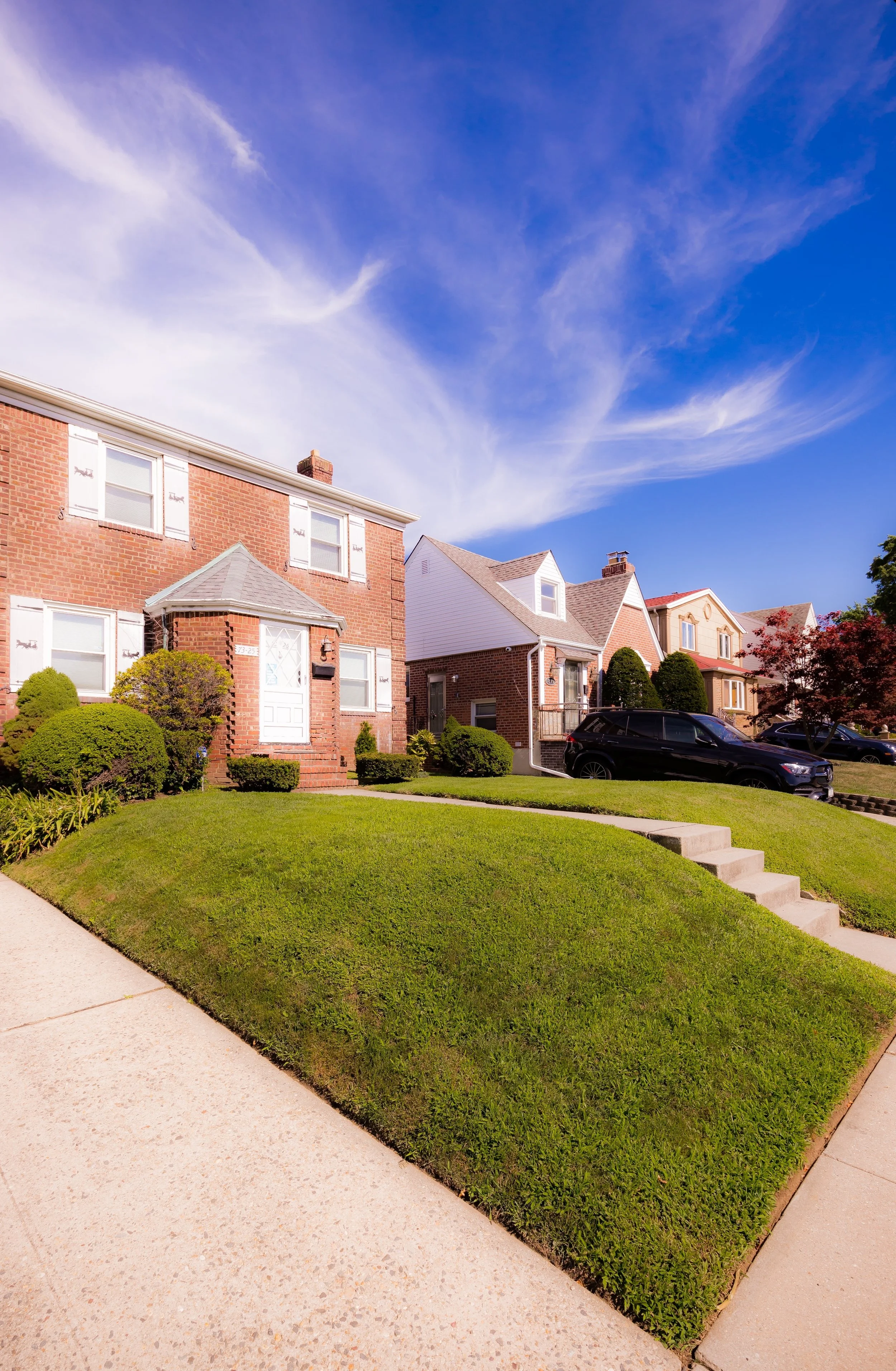 A row of suburban houses with front lawns, a blue sky with wispy clouds, and a parked black car.