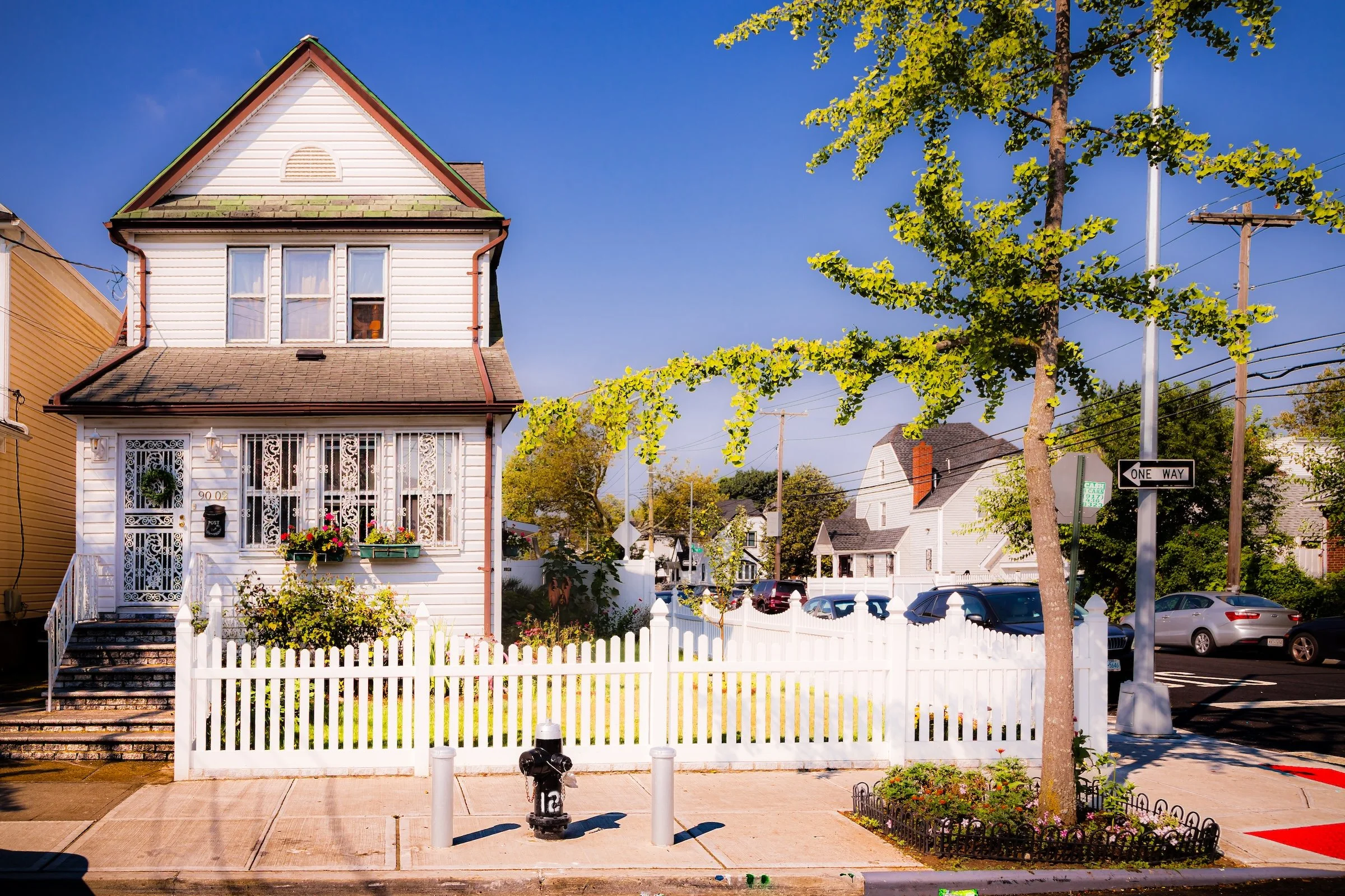 A brick house with a white section on the upper level, a front door with iron bars, and small bushes in front, flanked by two taller brick buildings under a clear blue sky.
