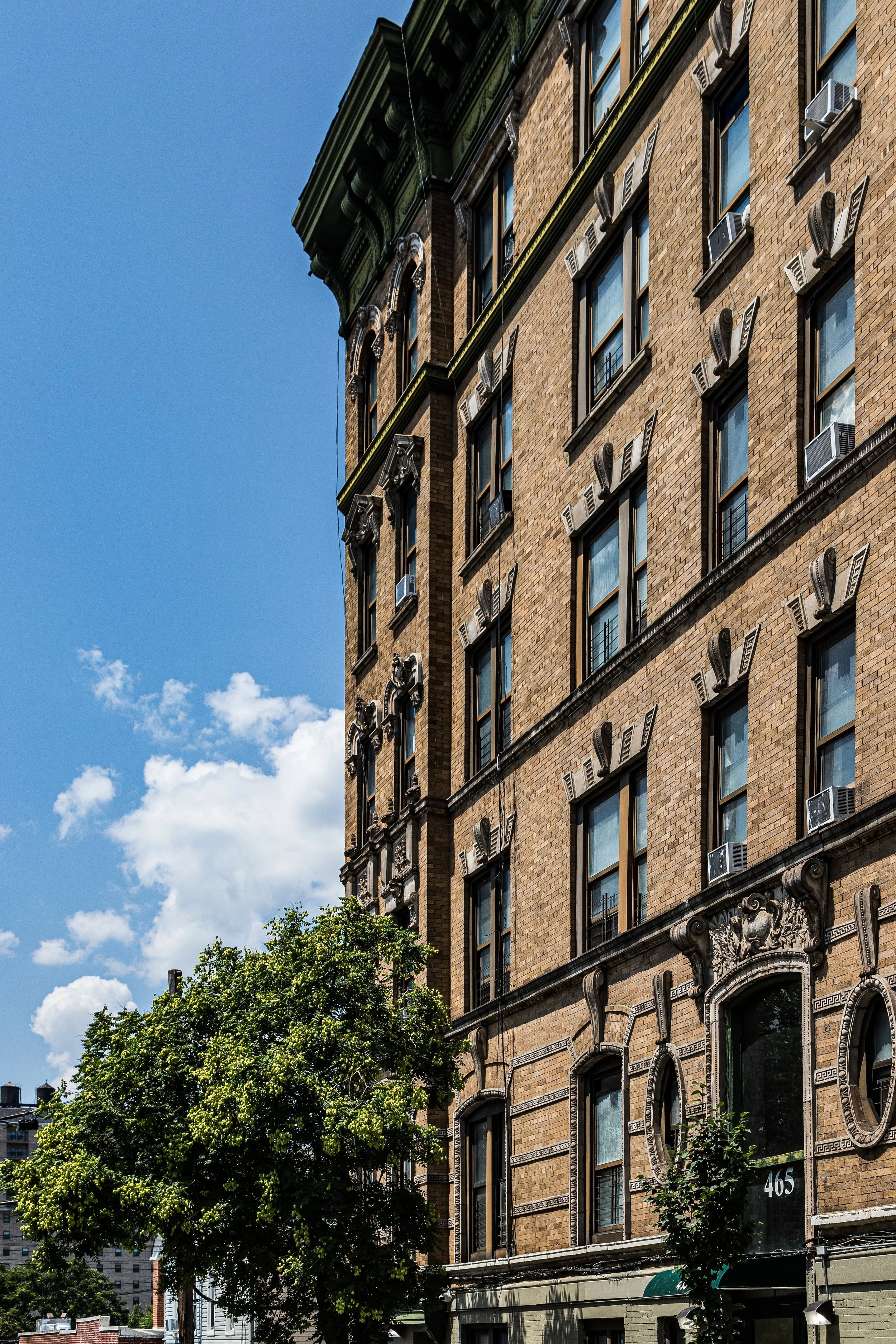 Tall brick building with decorative stonework and multiple windows, some with air conditioning units, next to a green tree, under a blue sky with scattered clouds.