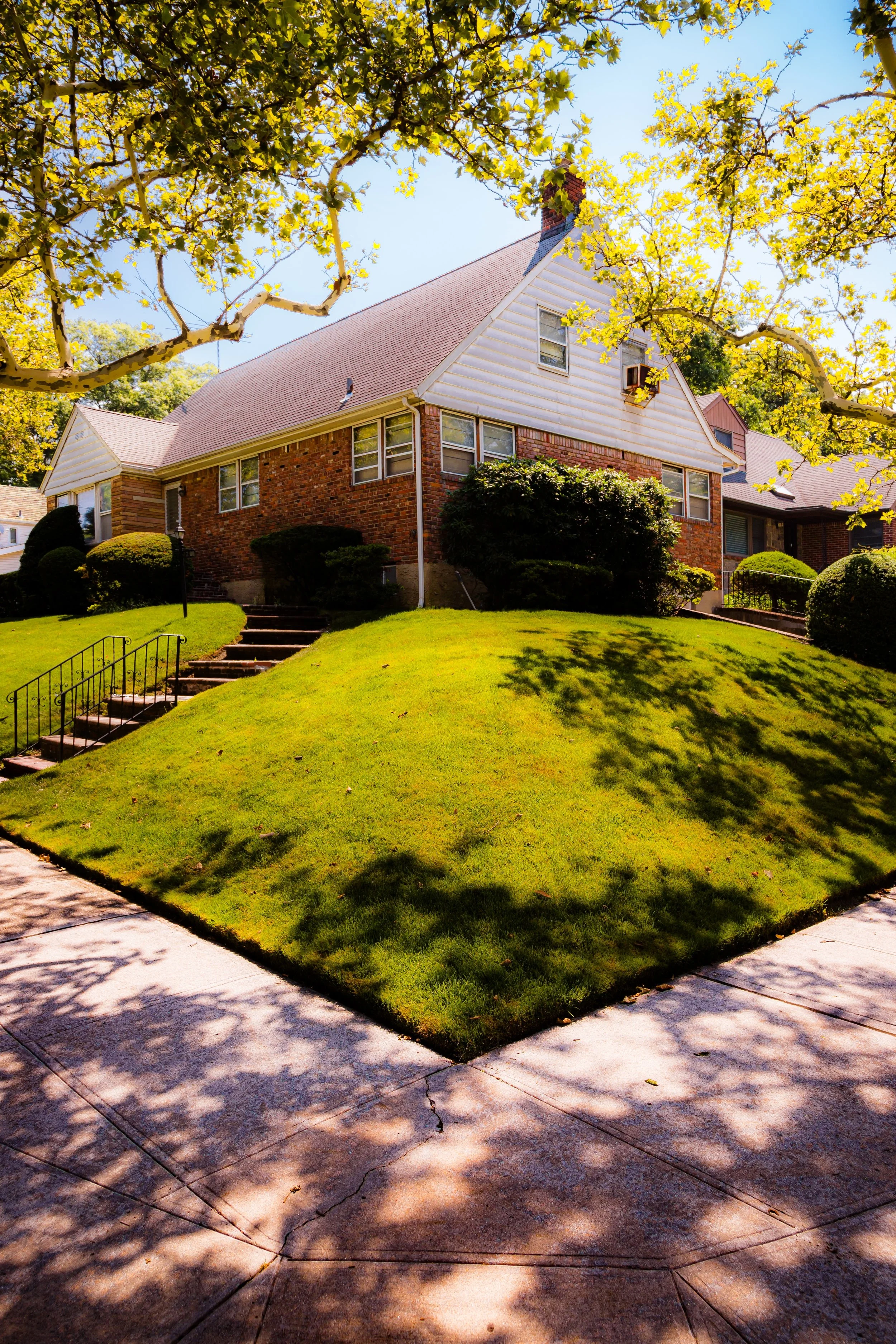 Front view of a brick and white house with a red roof, surrounded by green lawn and trees with yellow leaves, casting shadows on the sidewalk.