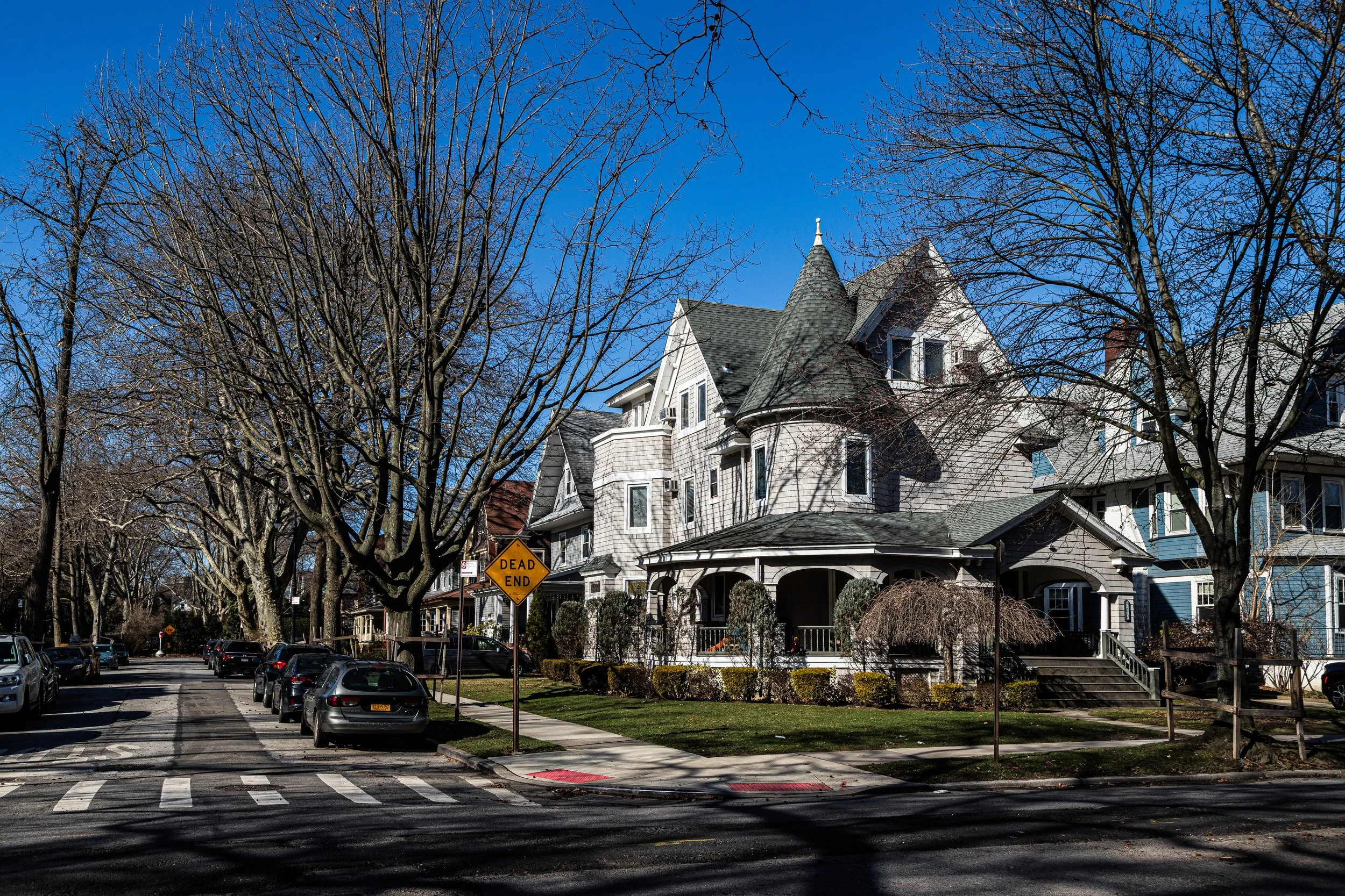 A street view with parked cars, leafless trees, and a large, Victorian-style house with a turret, porch, and stairs, under a clear blue sky.